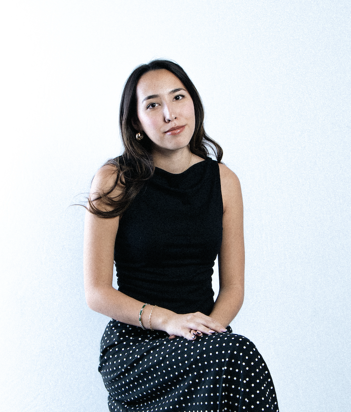 A woman with long dark hair wearing a black sleeveless top and a black skirt with white polka dots, sitting against a plain light-colored background.