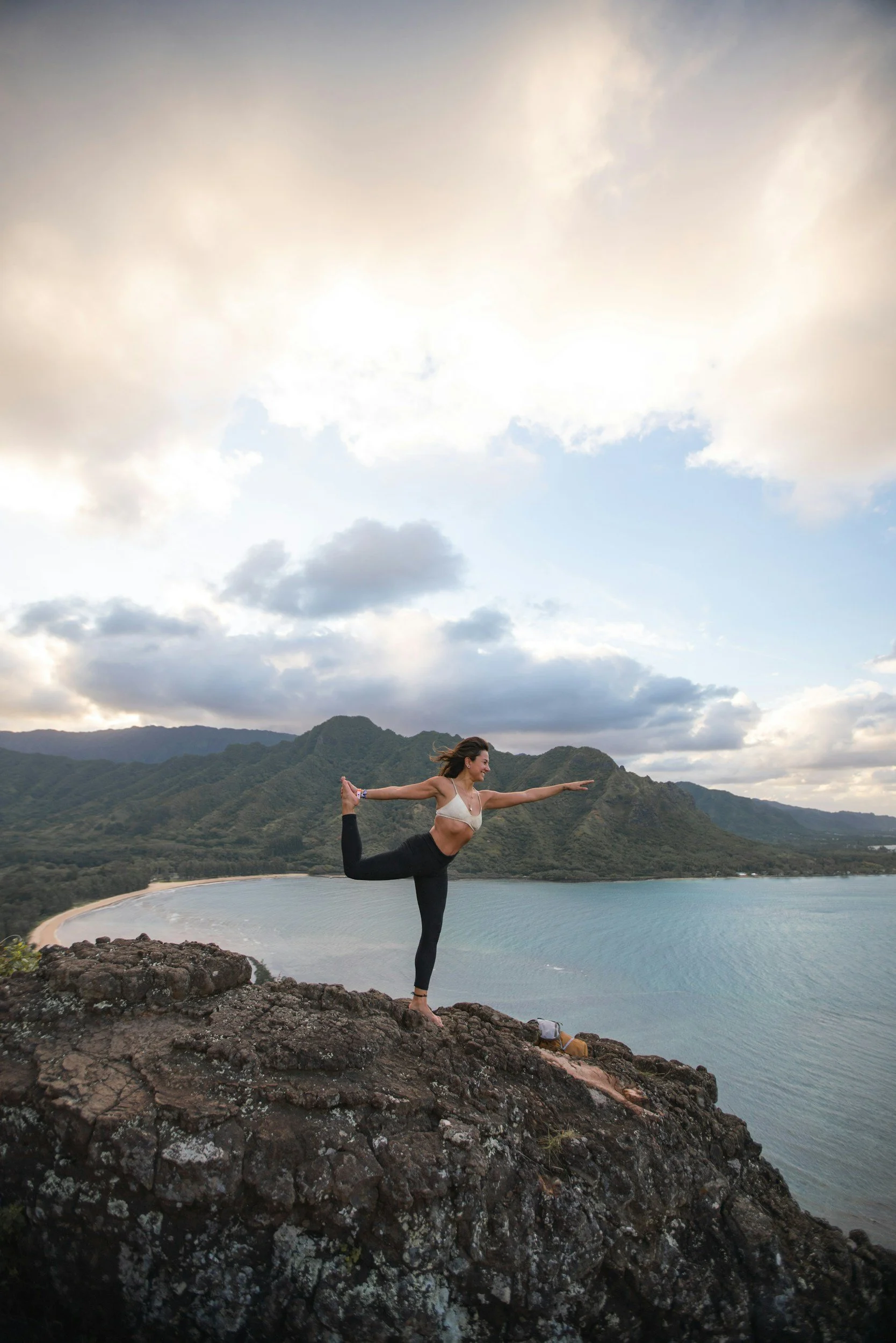 A woman practicing yoga on a rocky cliff overlooking the ocean with mountains and a cloudy sky in the background.