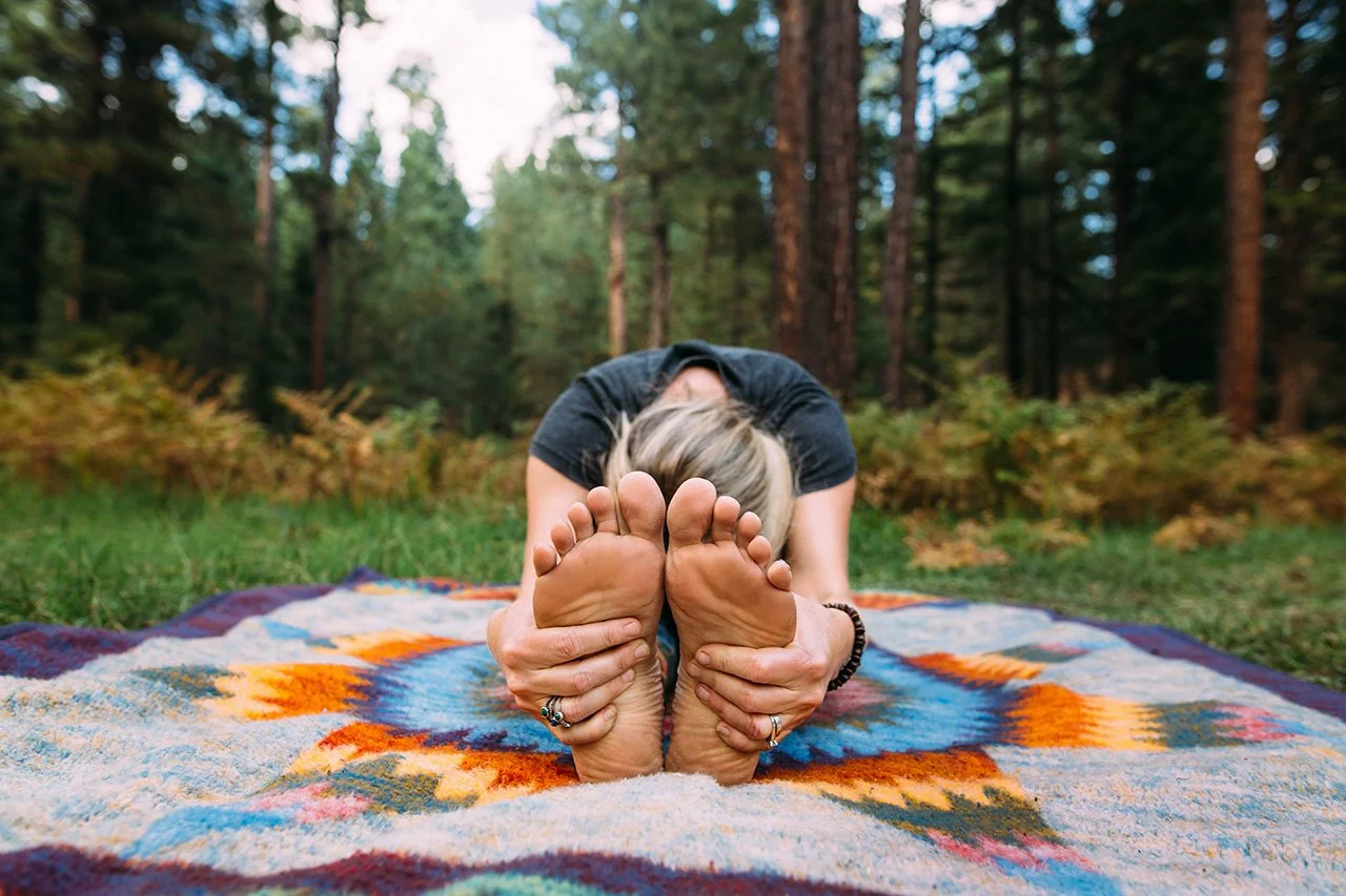 A woman practicing yoga outdoors on a colorful blanket in a forest, performing a seated forward bend with her feet touching and hands holding her soles.