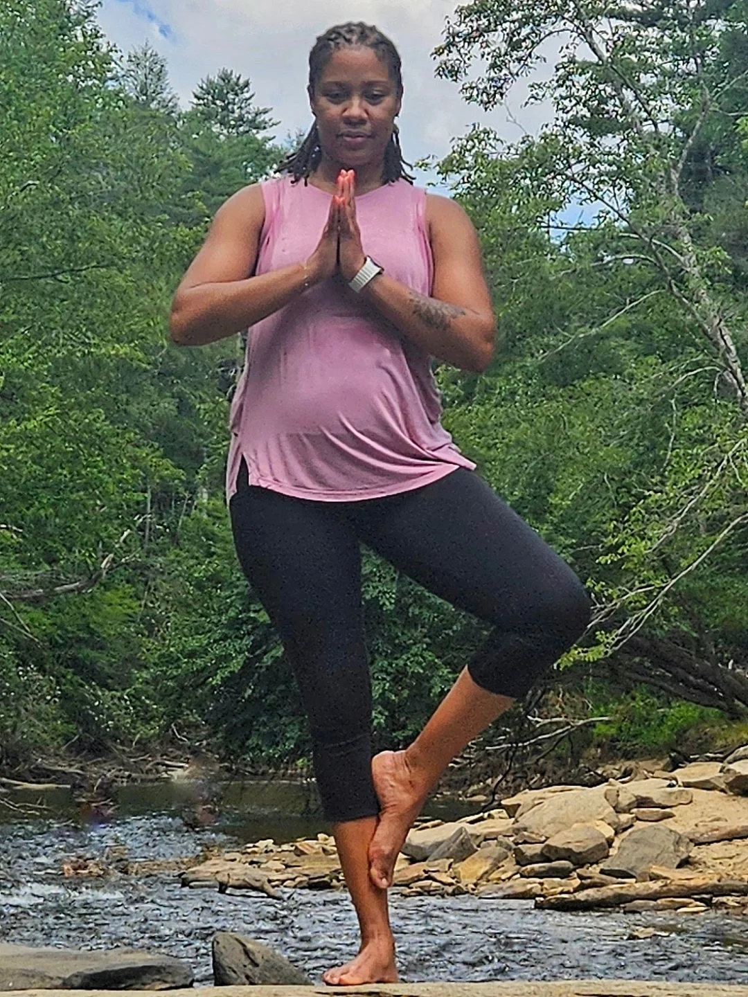 A woman practicing yoga outdoors near a stream, standing on one leg with her hands in a prayer position, surrounded by green trees.