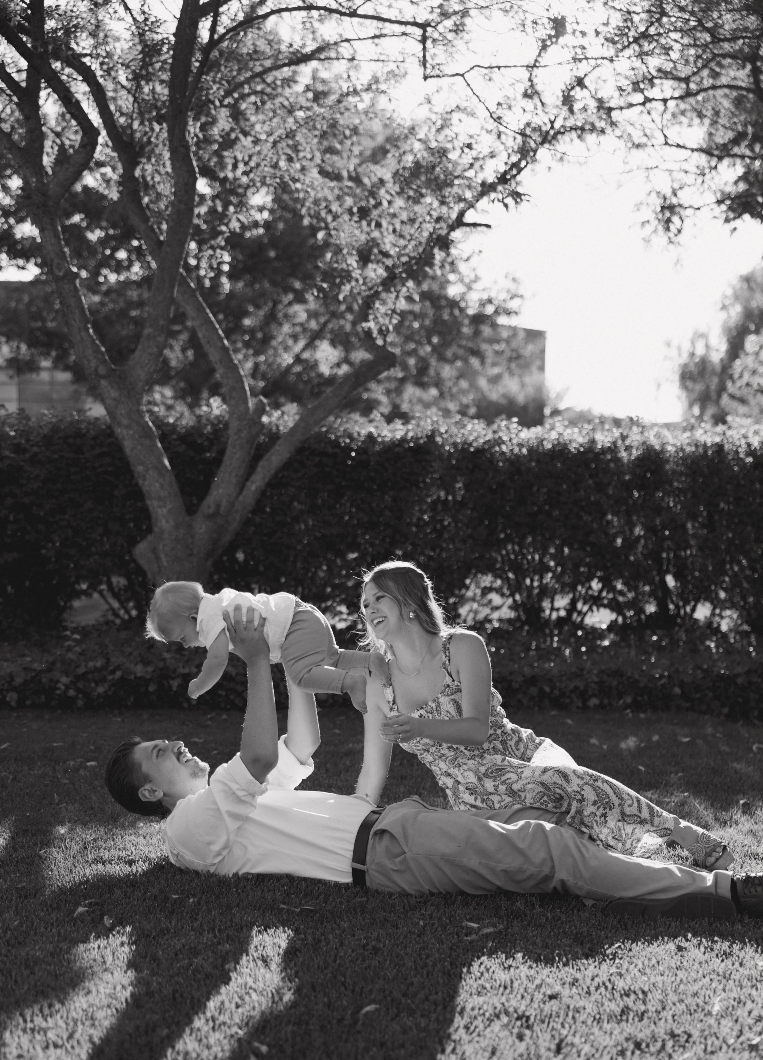 A family playing on the grass outdoors, with a man lying on his back, holding a child above him, while a woman sits nearby smiling, with trees and bushes in the background.