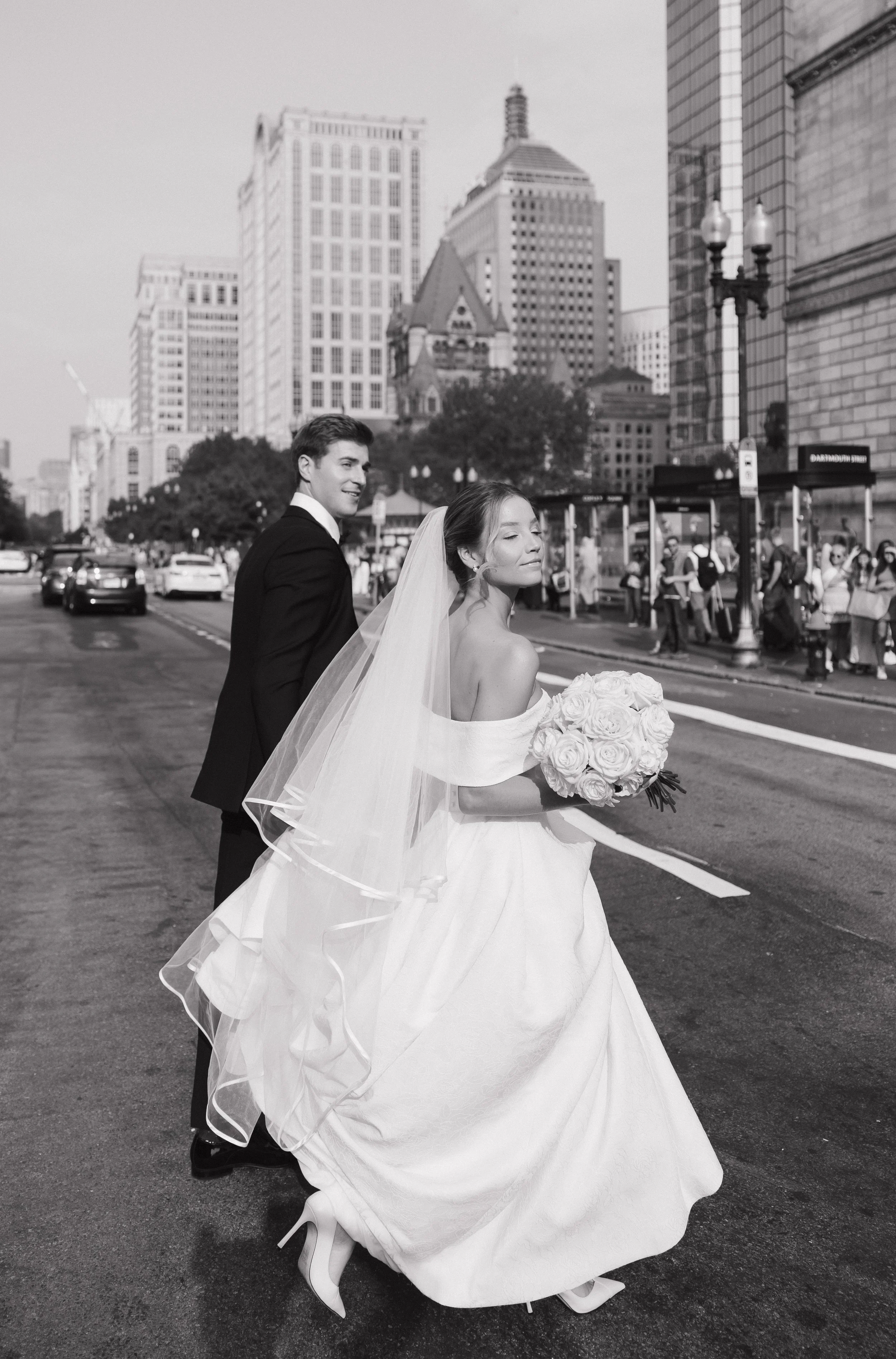 Black and white photo of a happy bride in a gown holding a bouquet of roses and a wedding veil, walking past a smiling groom in a suit. They are on a city street with tall buildings and people in the background.