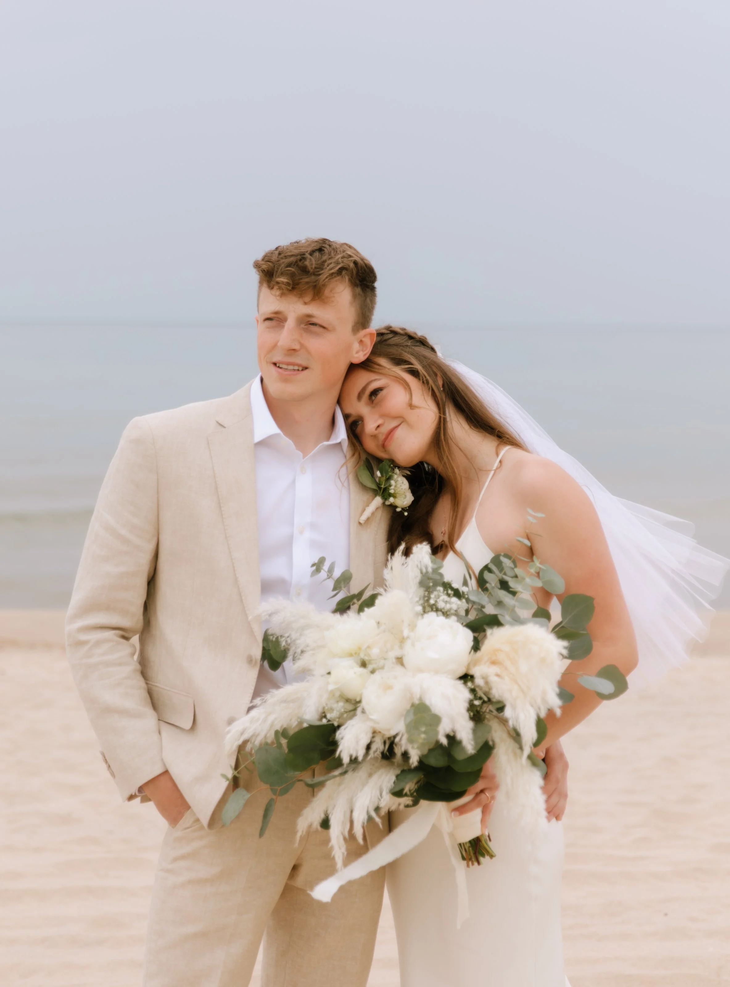 A bride and groom standing on a beach with the ocean in the background, holding a large bouquet of white flowers and green foliage.