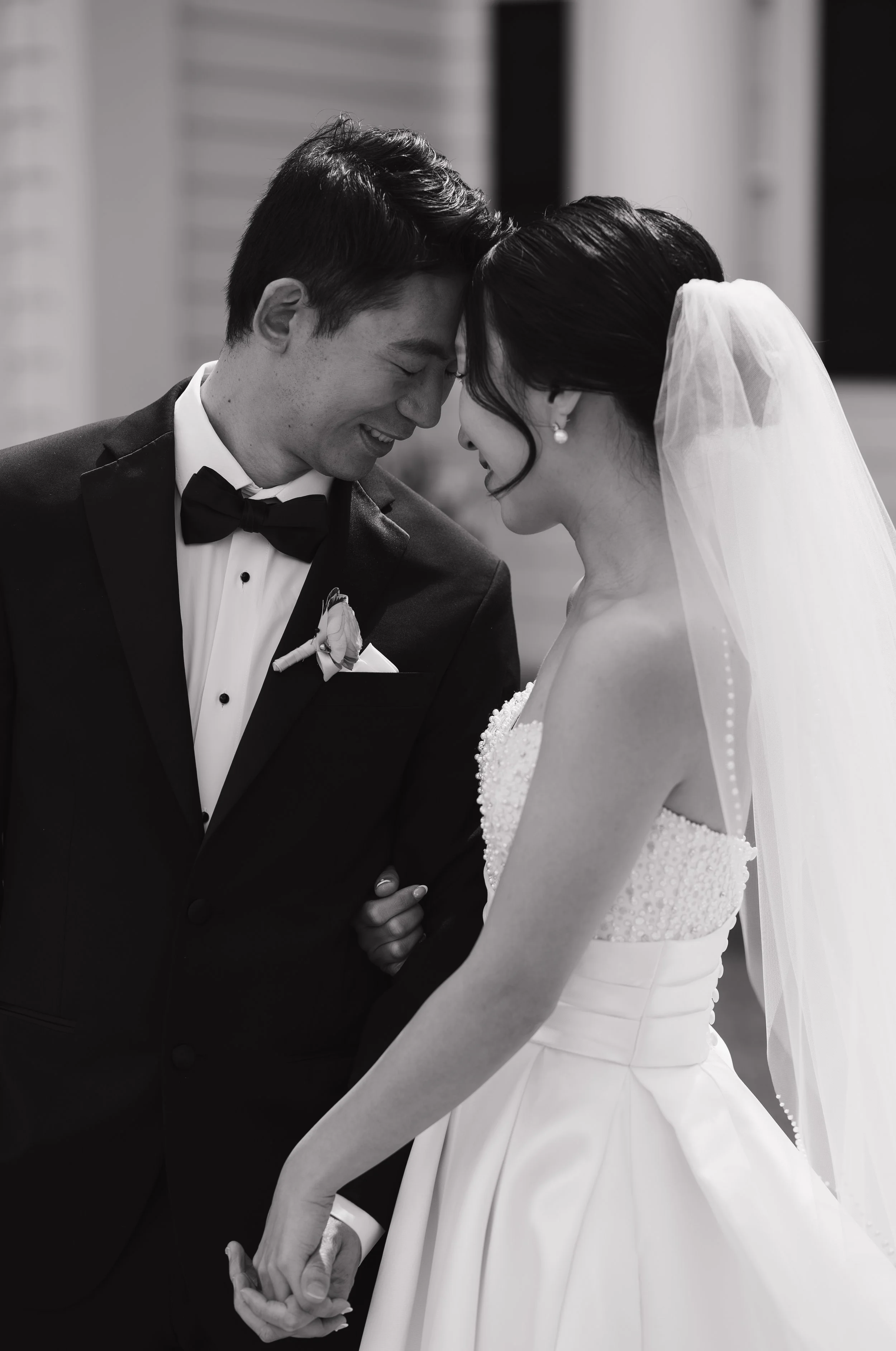 Black and white photo of a bride and groom in wedding attire holding hands and touching foreheads, smiling with closed eyes.