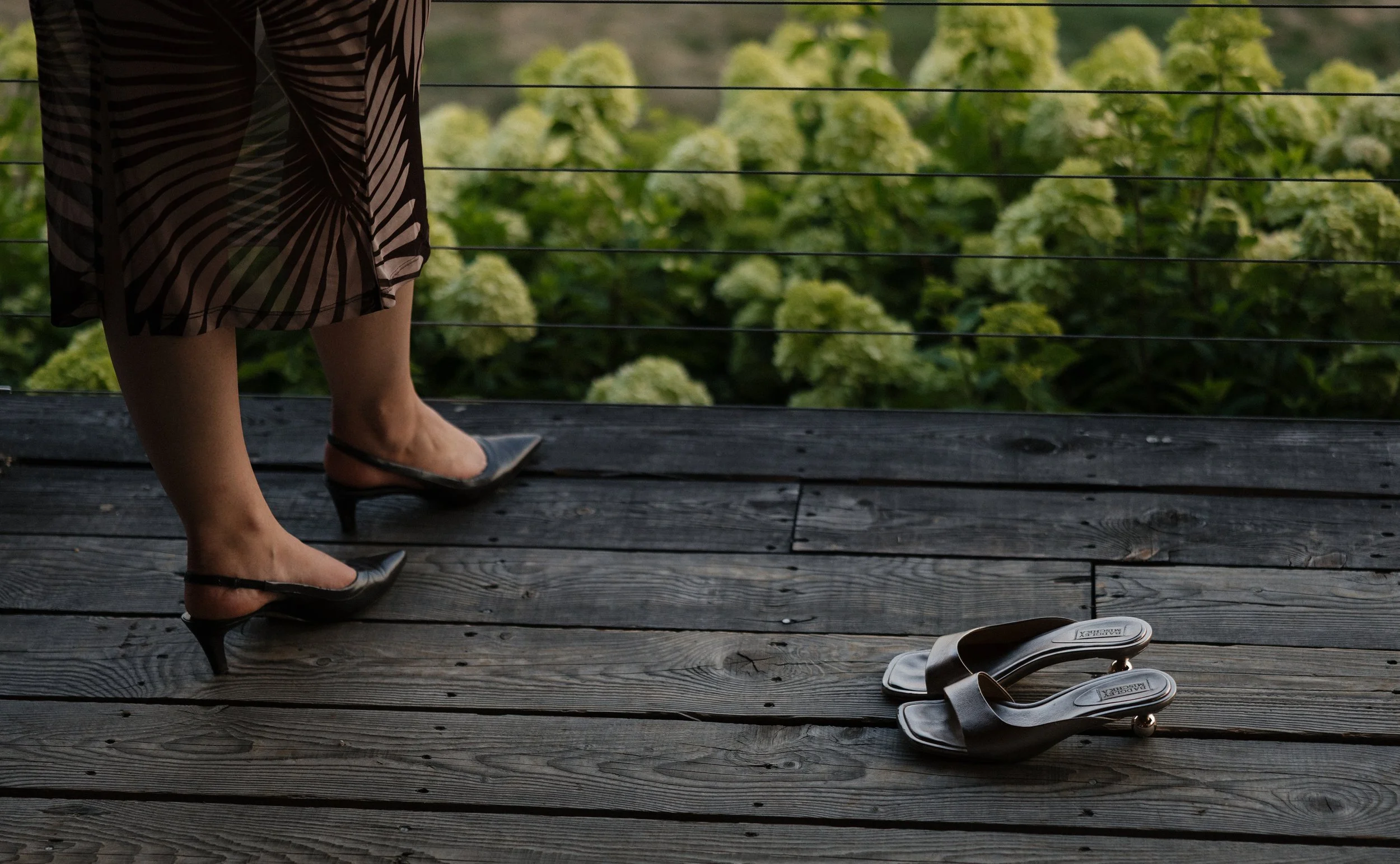 A person wearing a patterned skirt and black high heels standing on a wooden deck, with a white pair of shoes visible in the foreground, and a lush green shrubbery or garden in the background.