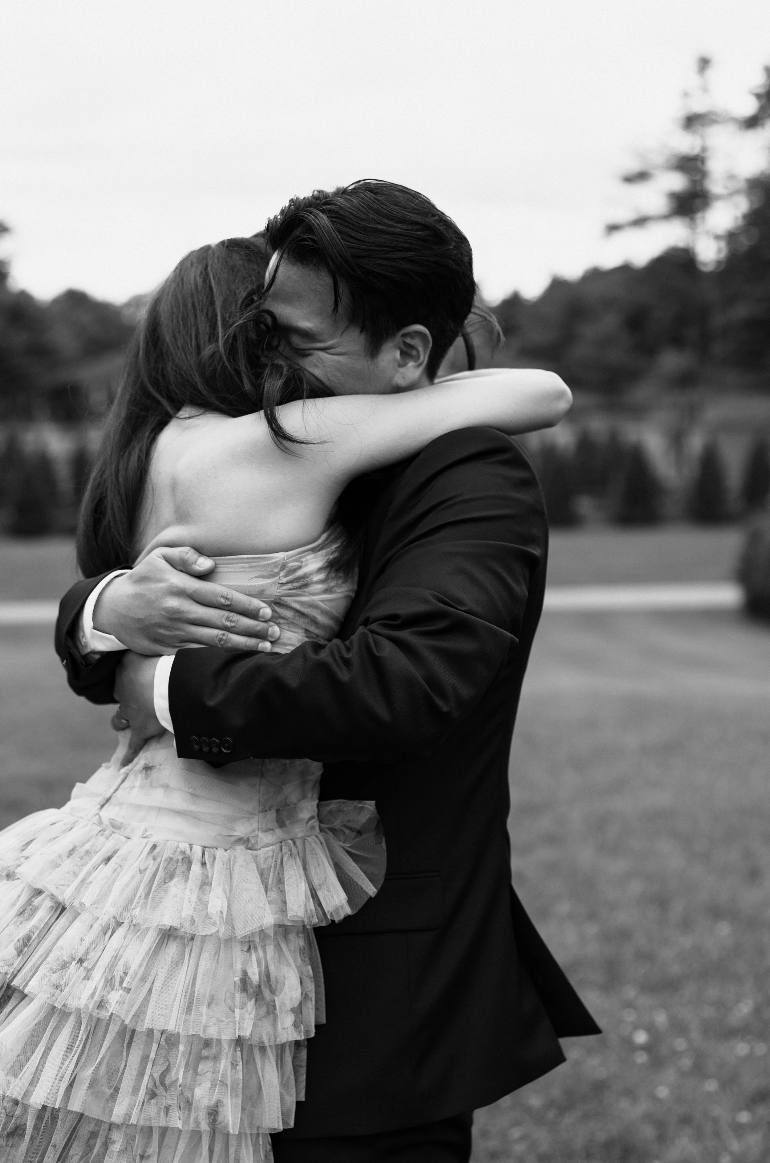 A man and a woman dressed in formal attire embracing outdoors, with the woman wearing a layered, ruffled dress and the man in a suit, both smiling with their faces touching.