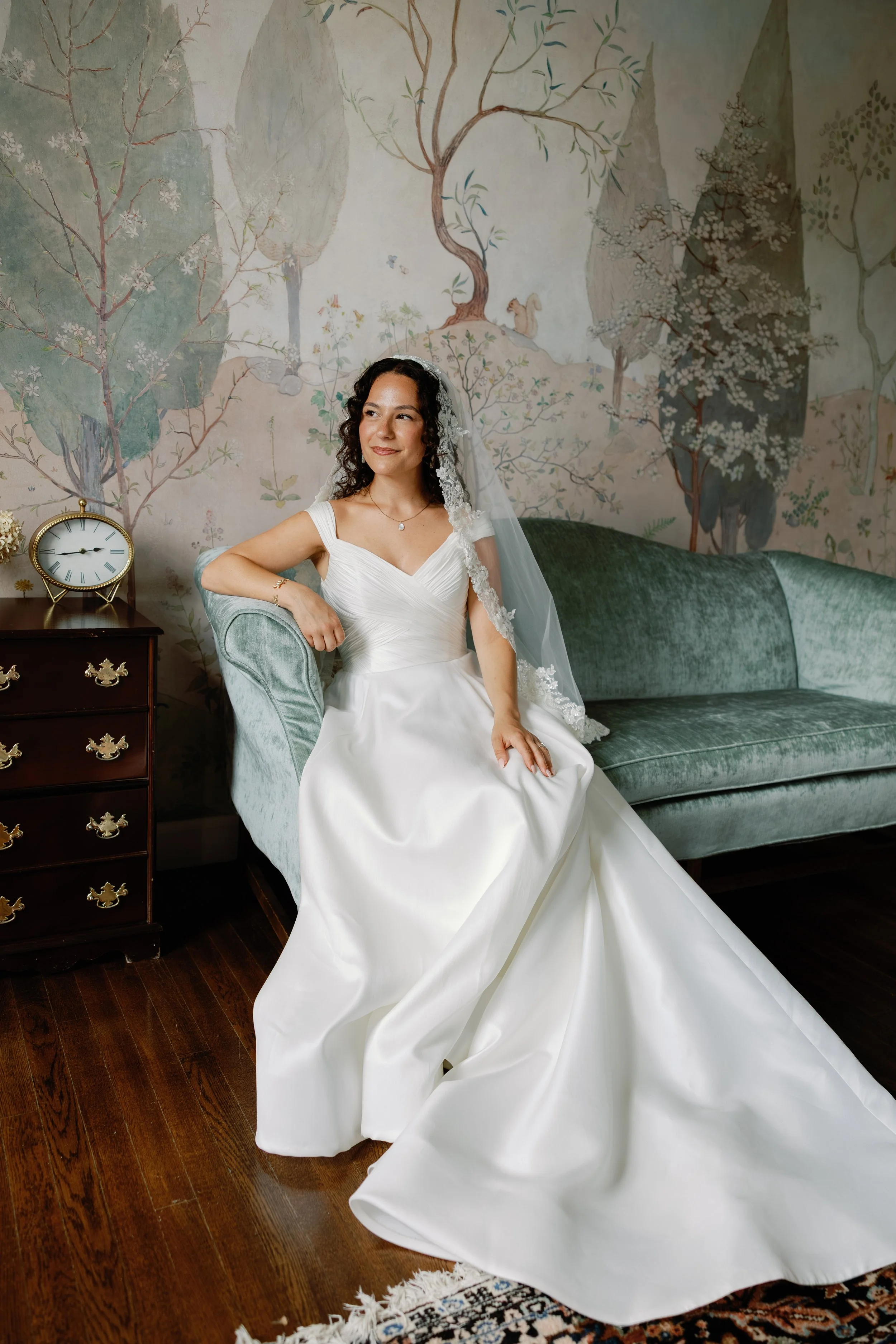 A woman in a white wedding dress sitting on a vintage green velvet sofa in a room with a painted mural of trees and mountains on the wall behind her, and a small wooden side table with a clock and flowers to her left.