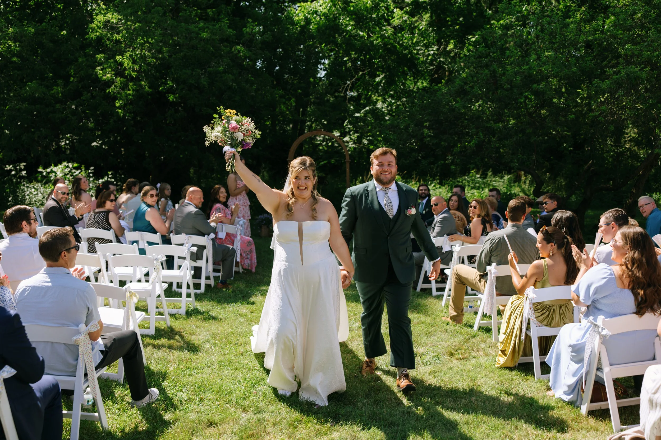 A bride and groom walk hand-in-hand down the aisle at an outdoor wedding, the bride holding a bouquet of flowers and smiling, while guests seated in white chairs applaud and celebrate.