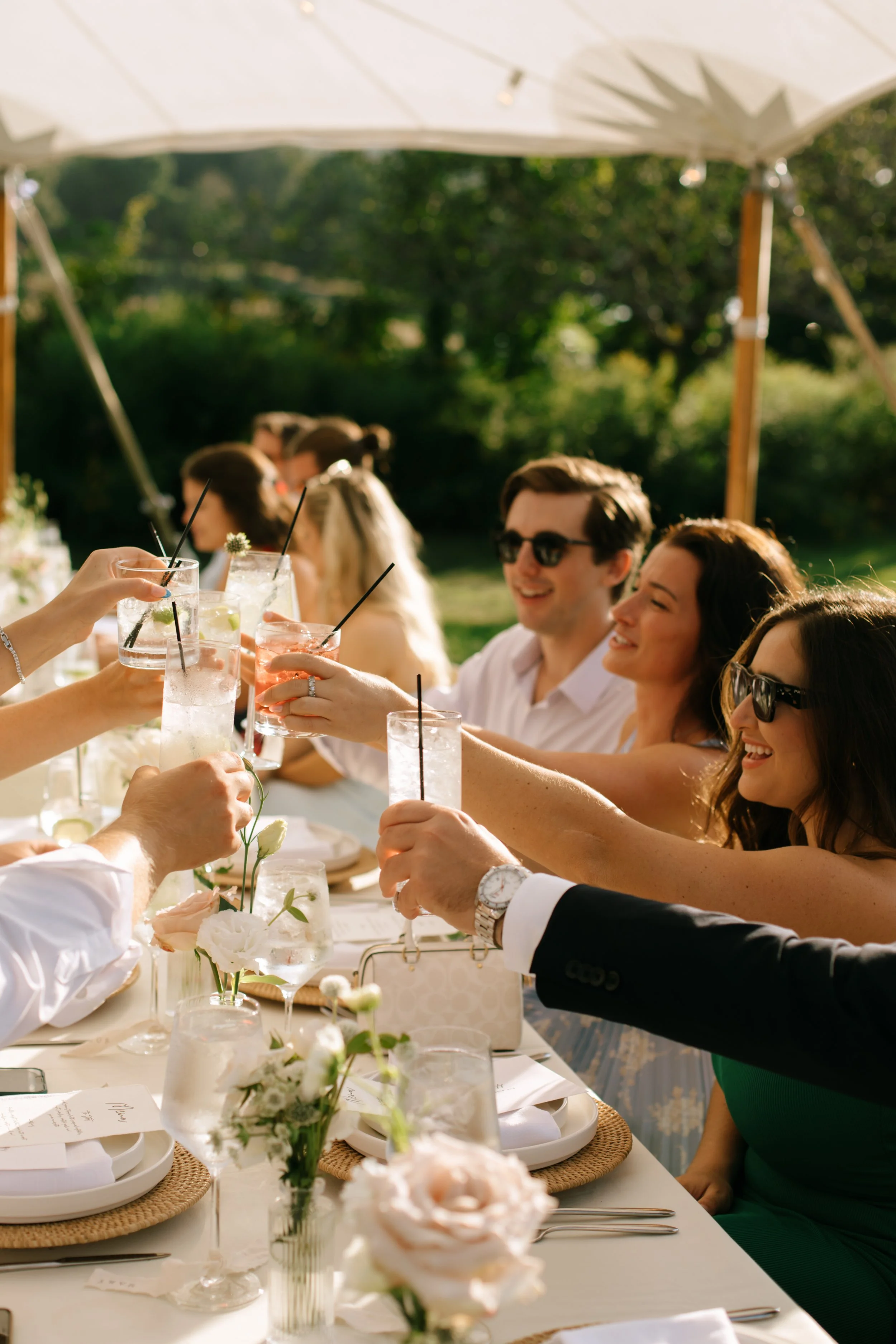 People raising glasses with drinks at an outdoor celebration, smiling and enjoying each other's company under a white canopy.