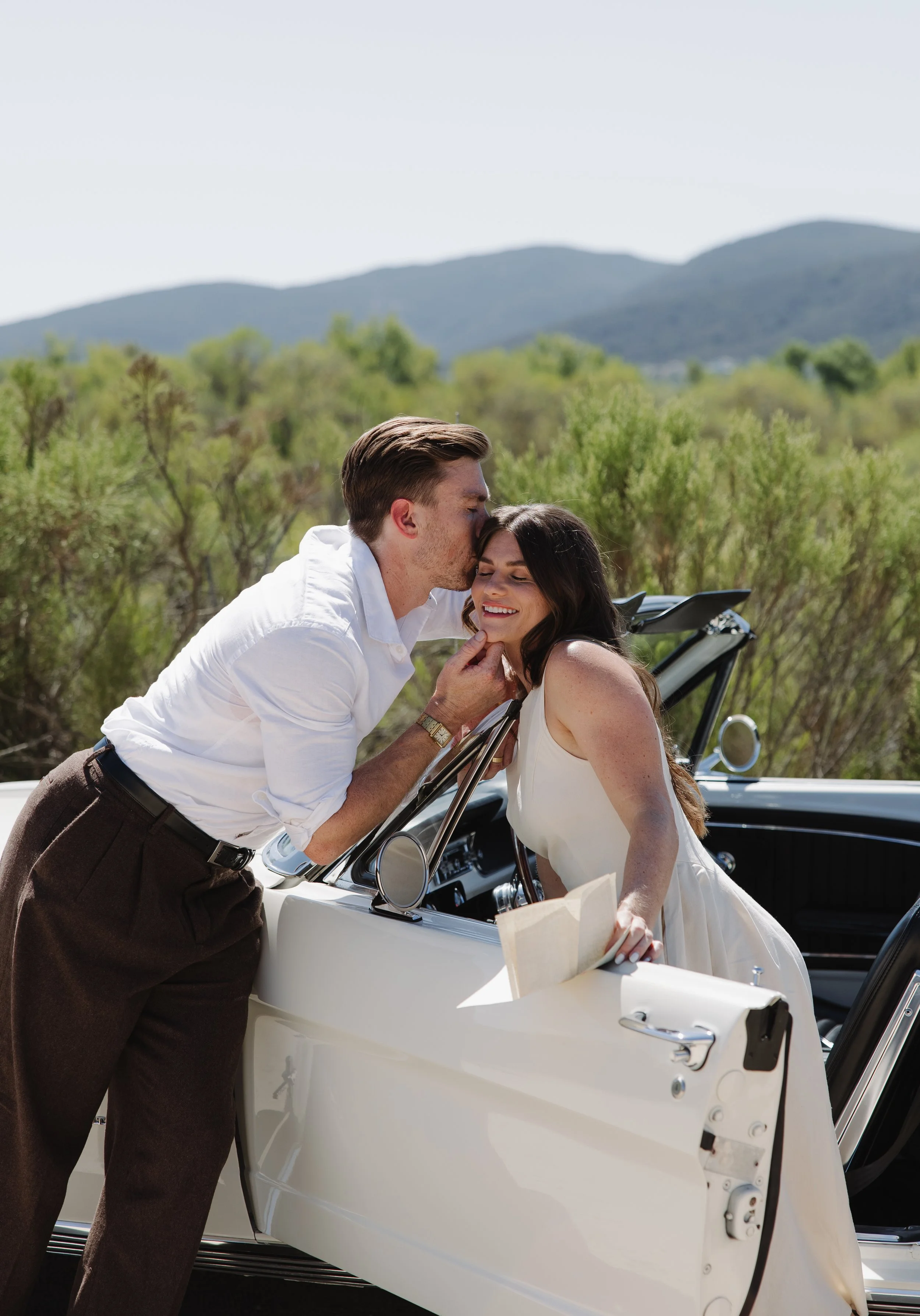 A man in a white shirt and dark pants kisses a woman in a white dress on the forehead while leaning over a classic white convertible car, with a scenic landscape of trees and mountains in the background.