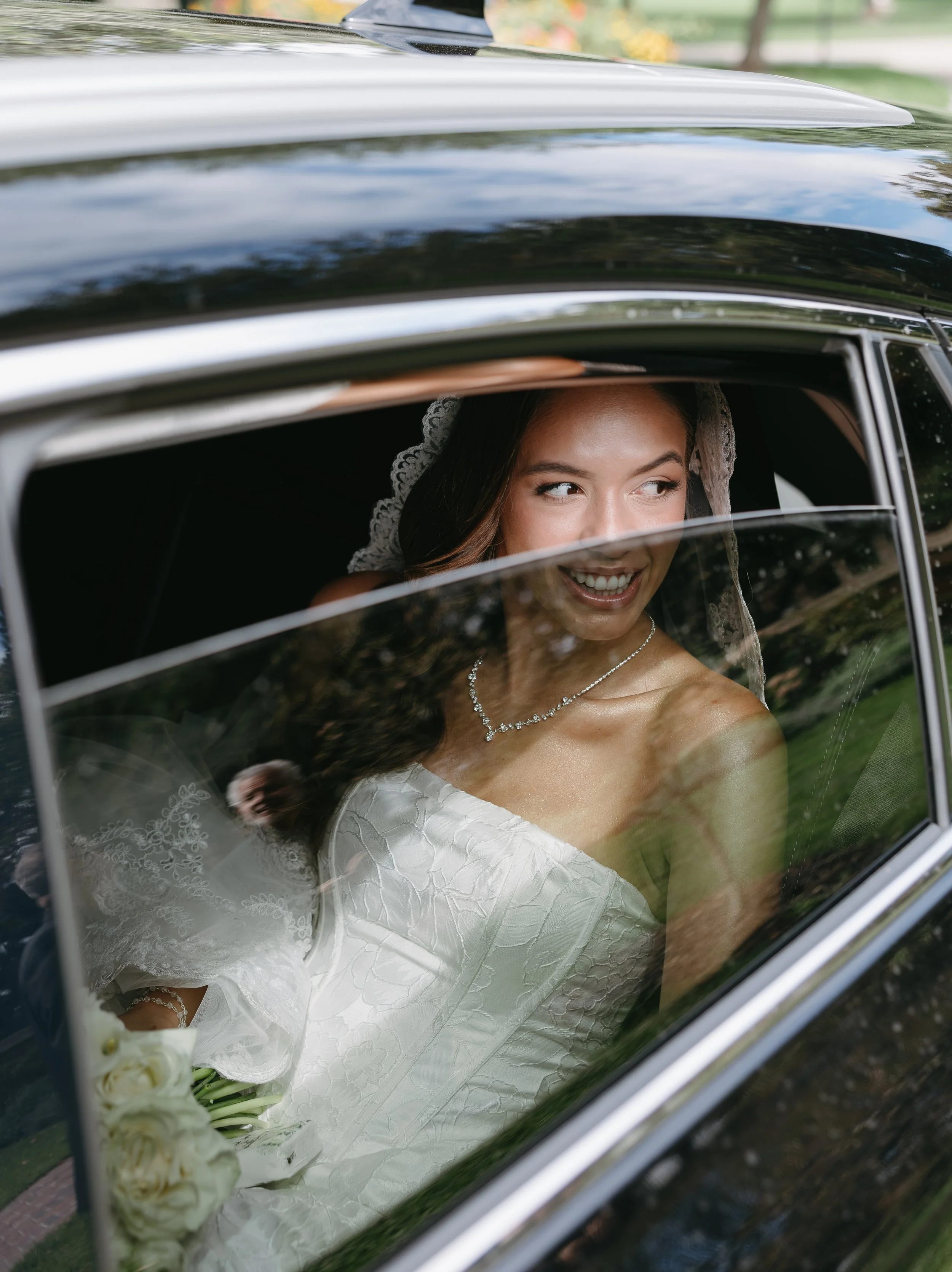 A bride with a lace veil and diamond necklace sitting in a black car, smiling and looking out the window.