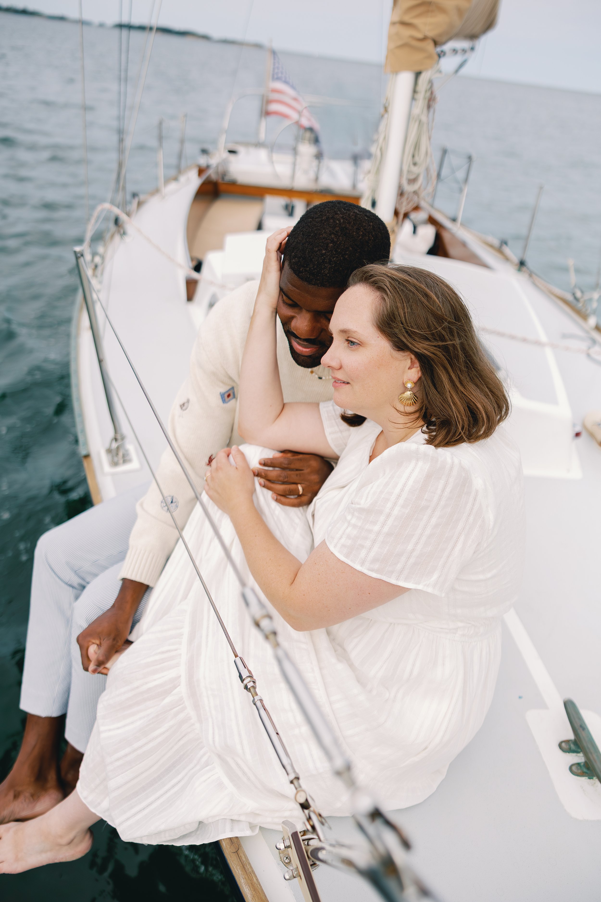 A man and a woman sitting closely together on the deck of a sailboat, with the woman leaning her head on the man's shoulder, surrounded by water.