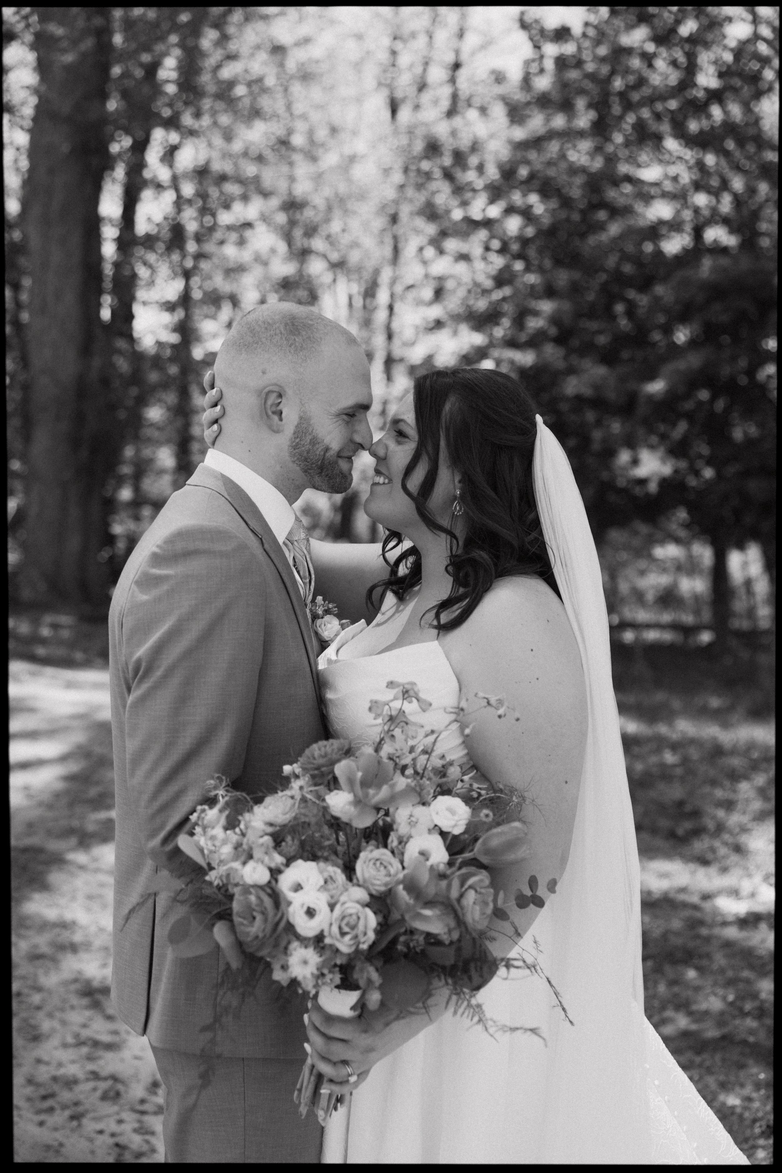 Black and white photo of a bride and groom touching foreheads and smiling, standing outdoors with trees in the background. The bride is holding a large bouquet of flowers and wearing a wedding dress, while the groom is in a suit.