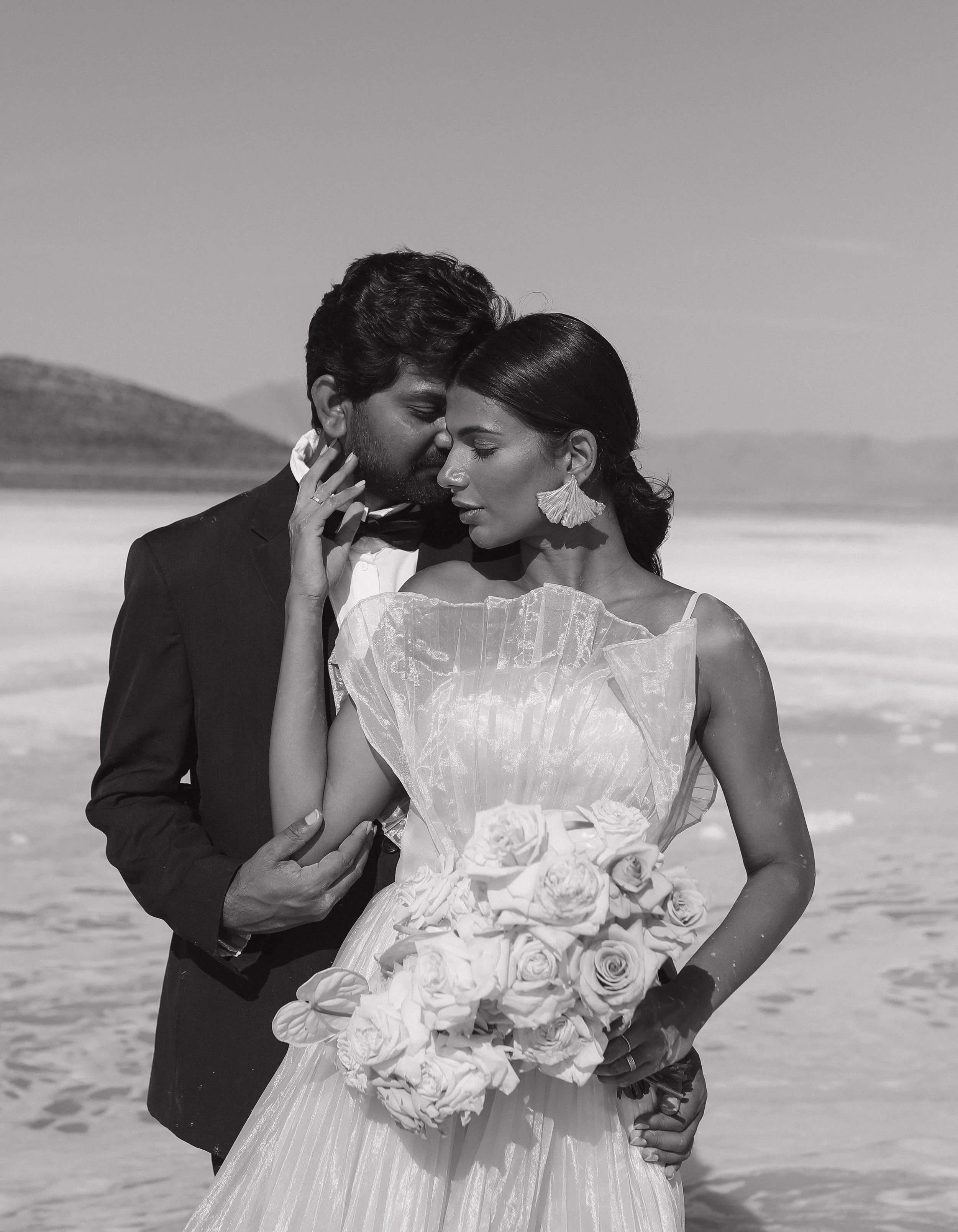 A black-and-white photo of a couple dressed in formal attire, embracing closely on a beach with dunes in the background. The man is wearing a tuxedo and the woman is in a wedding dress holding a bouquet of roses.