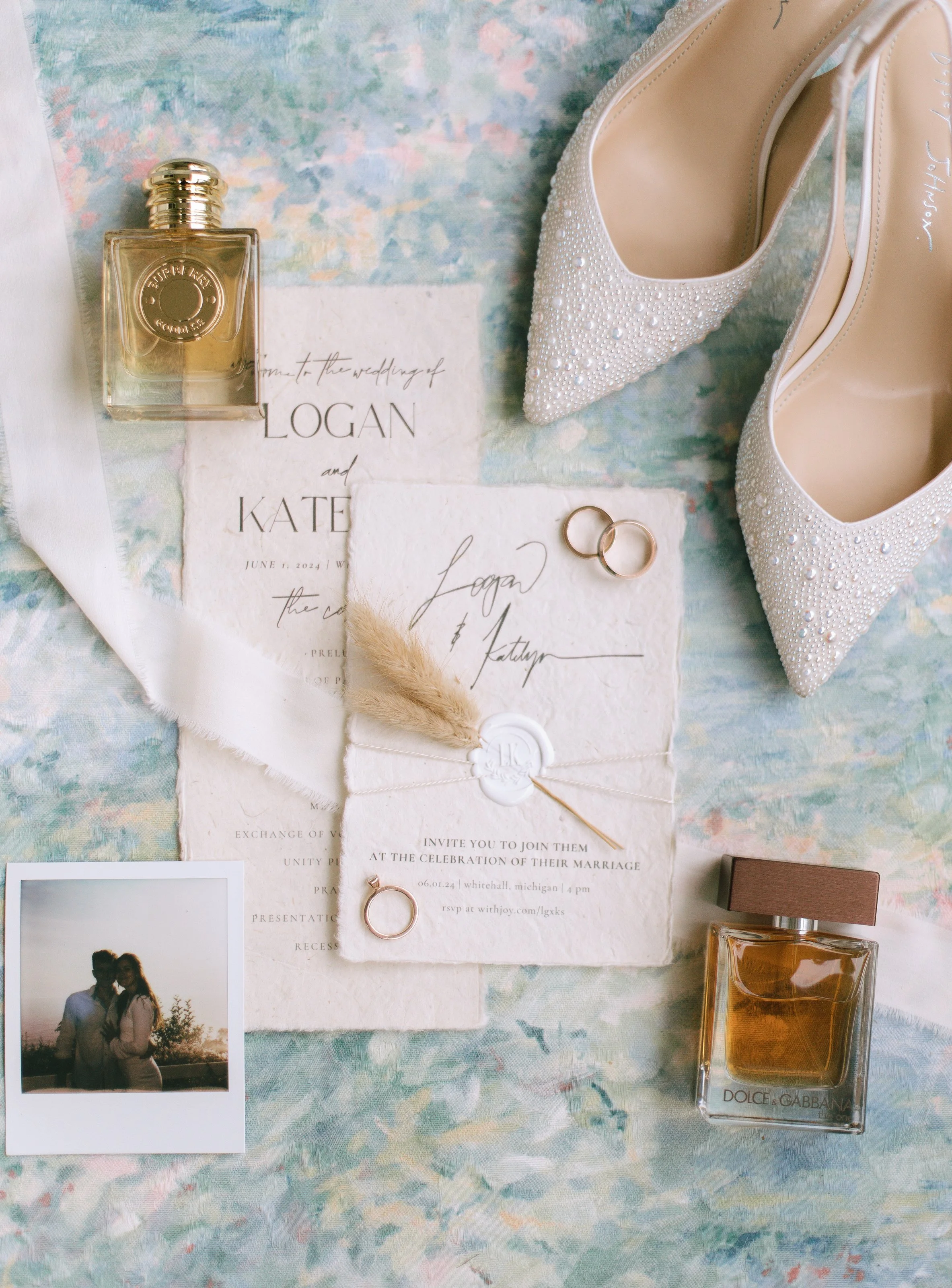 Wedding invitation, perfume bottle, pearl embellished shoes, wedding rings, and a small photo of a couple, all laid out on a pastel watercolor fabric background.