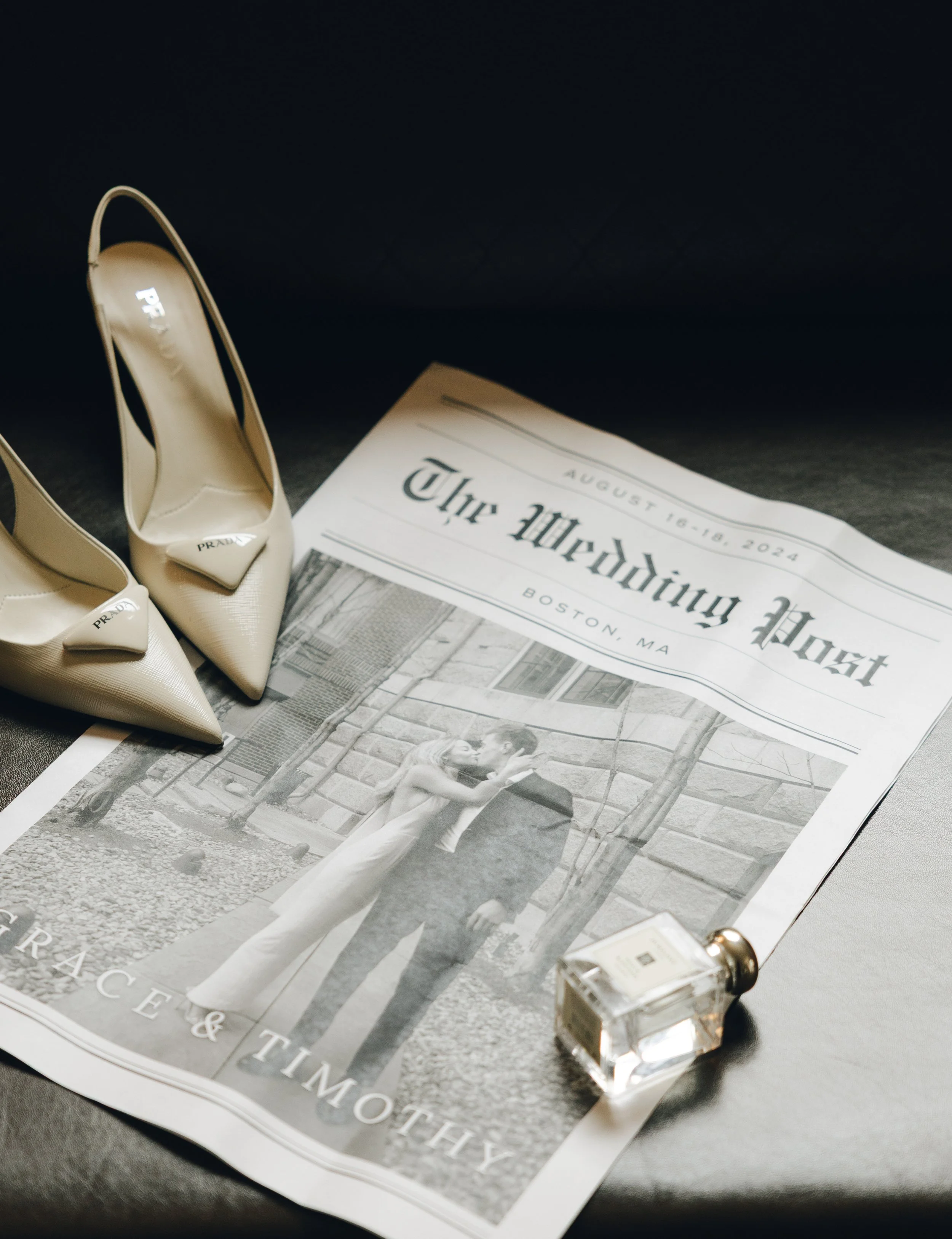 A pair of beige Prada high-heeled shoes with pointed toes and ankle straps resting on a magazine, next to a small transparent perfume bottle with gold accents.