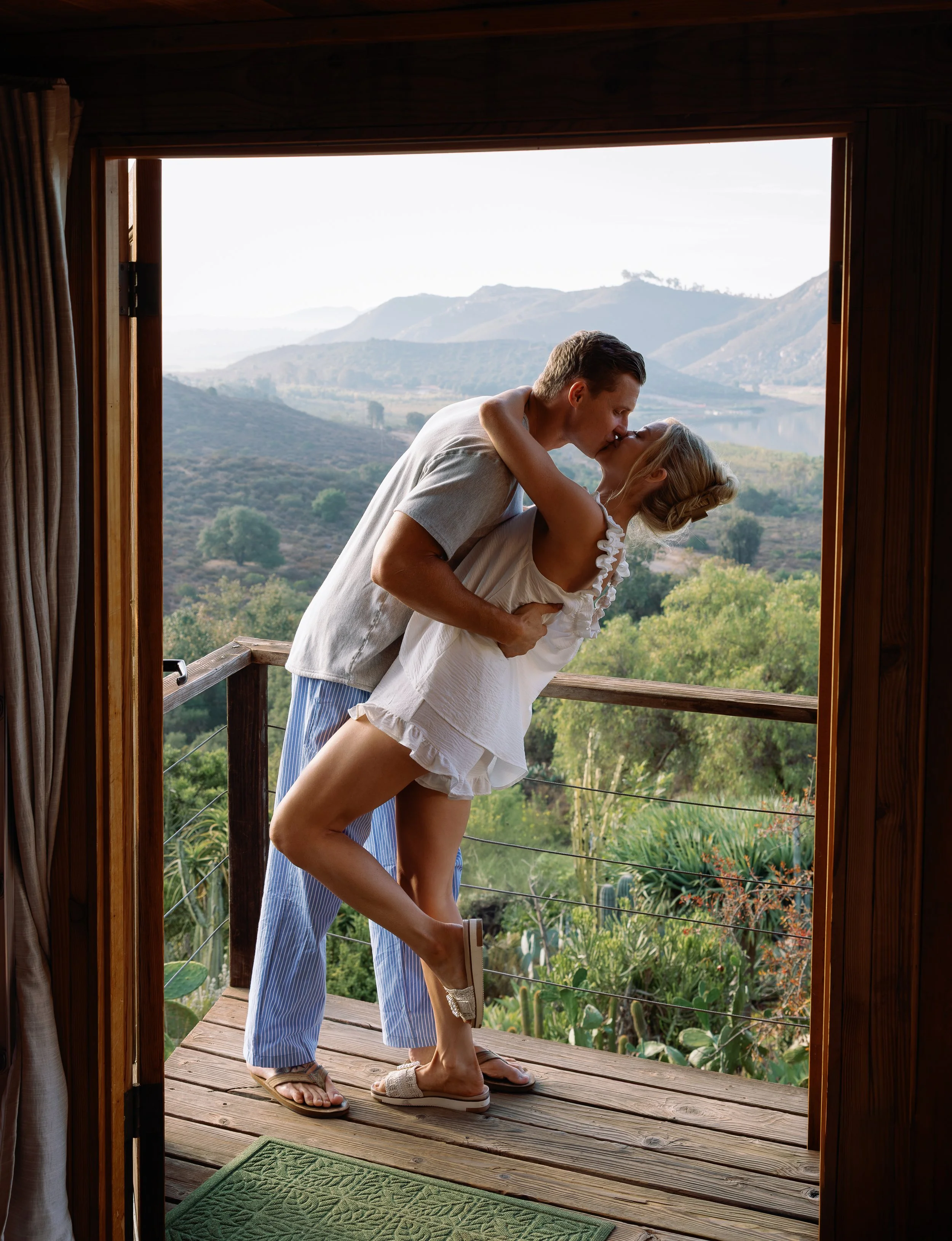 A couple kissing on a wooden balcony overlooking a lush, mountainous landscape with a lake in the distance, during daytime.