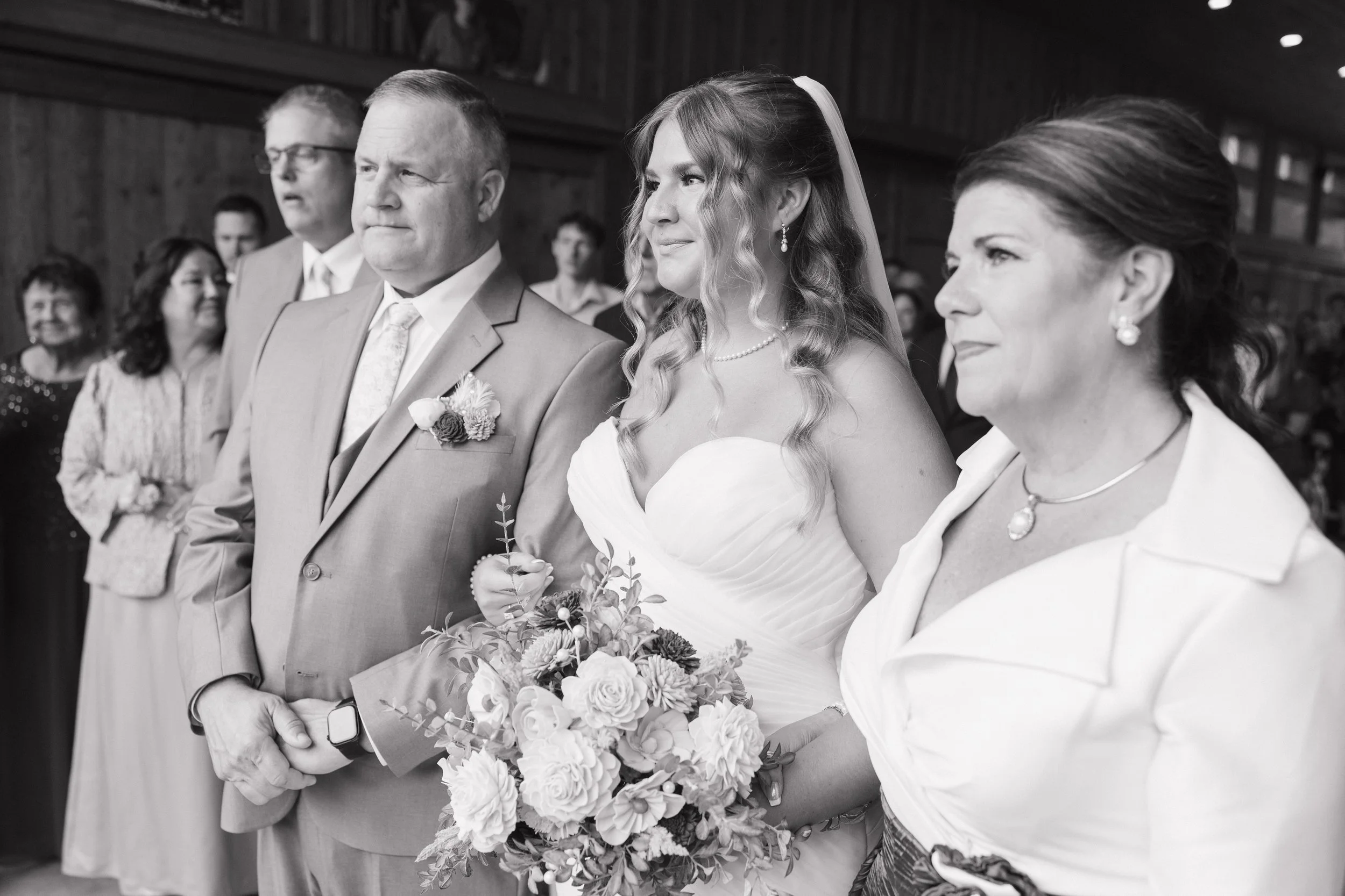 Black and white photo of a wedding ceremony, focusing on the bride and groom standing side by side holding a bouquet of flowers, surrounded by guests.