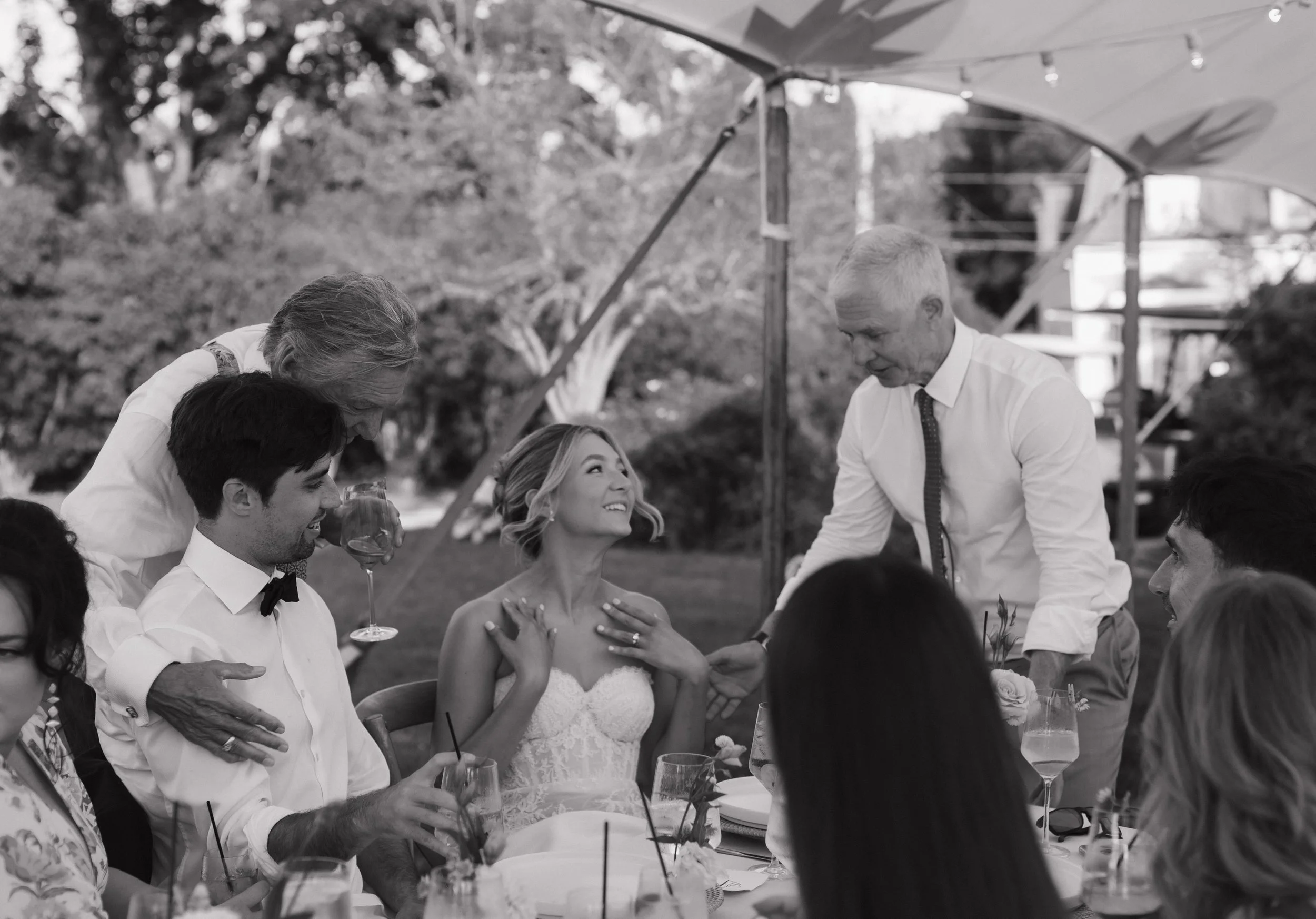 A black-and-white photo of a wedding celebration outdoors. The bride is in a strapless white dress, smiling and looking at an older man leaning over her. The groom, in a tuxedo with a bowtie, has his eyes closed and is smiling. Several guests are gat