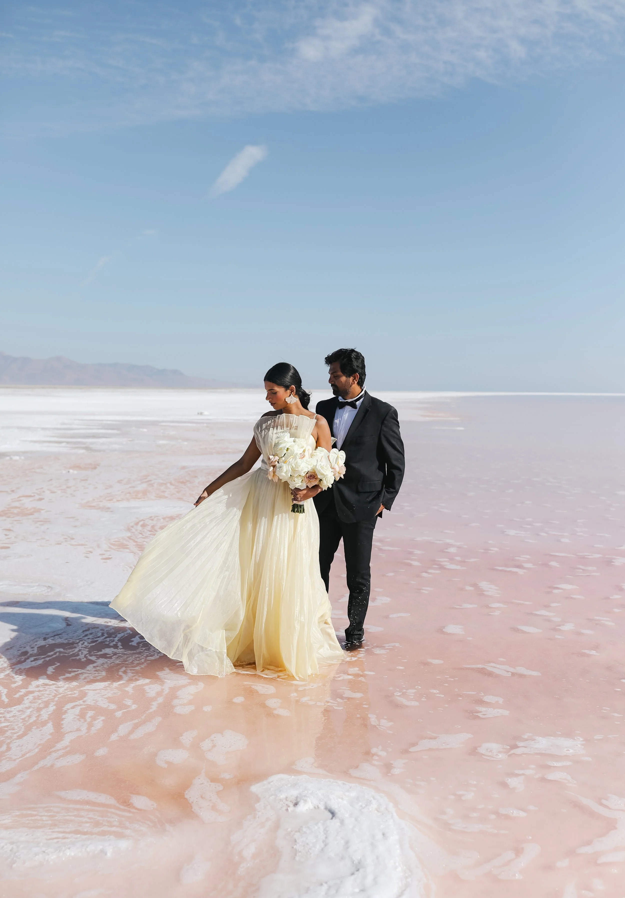 A couple dressed in wedding attire walking in shallow pink saltwater with a clear blue sky overhead.