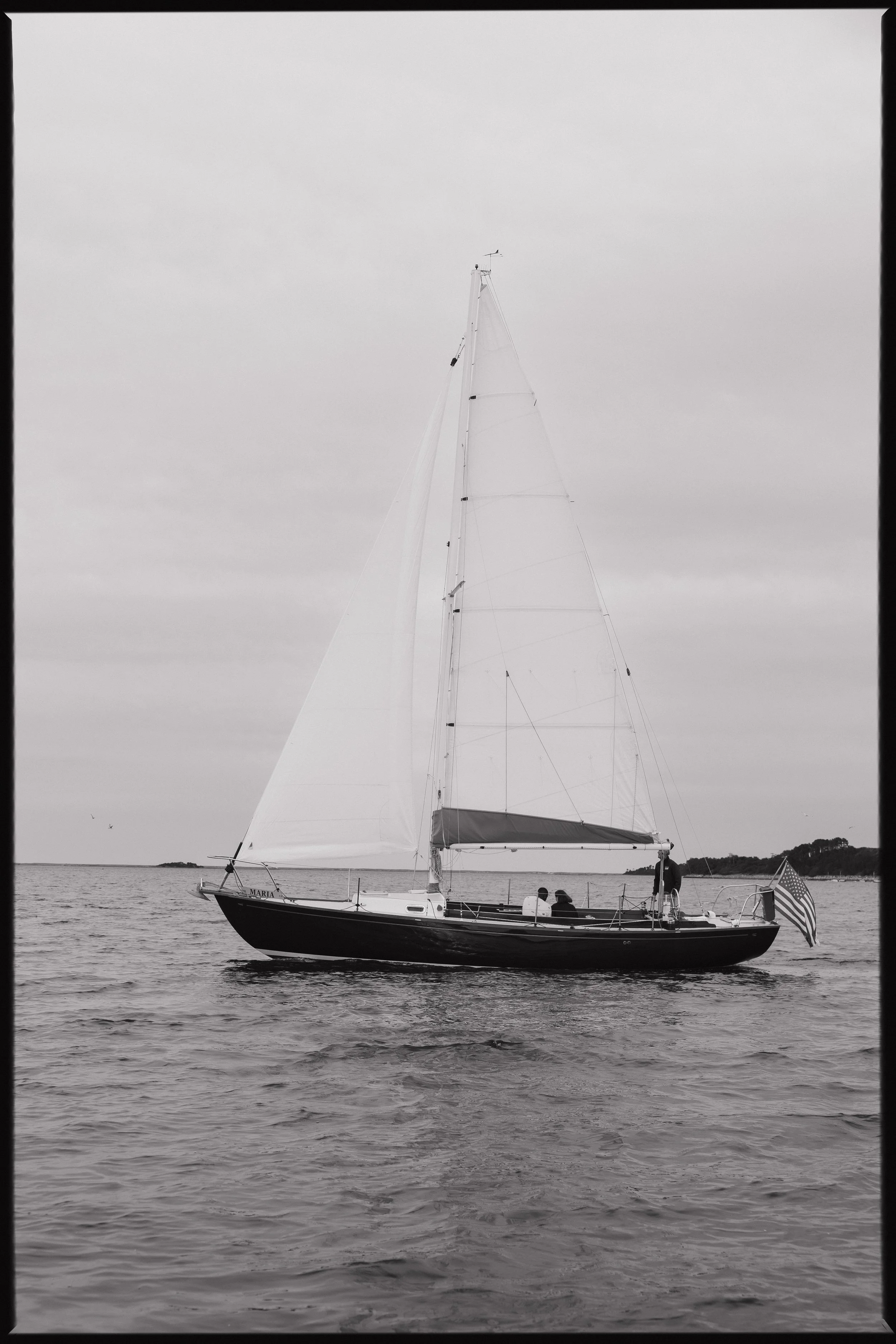 Black and white photograph of a sailboat with its sails up on calm water, with a small landmass or island in the background, and three people on board near the stern, with an American flag at the stern.