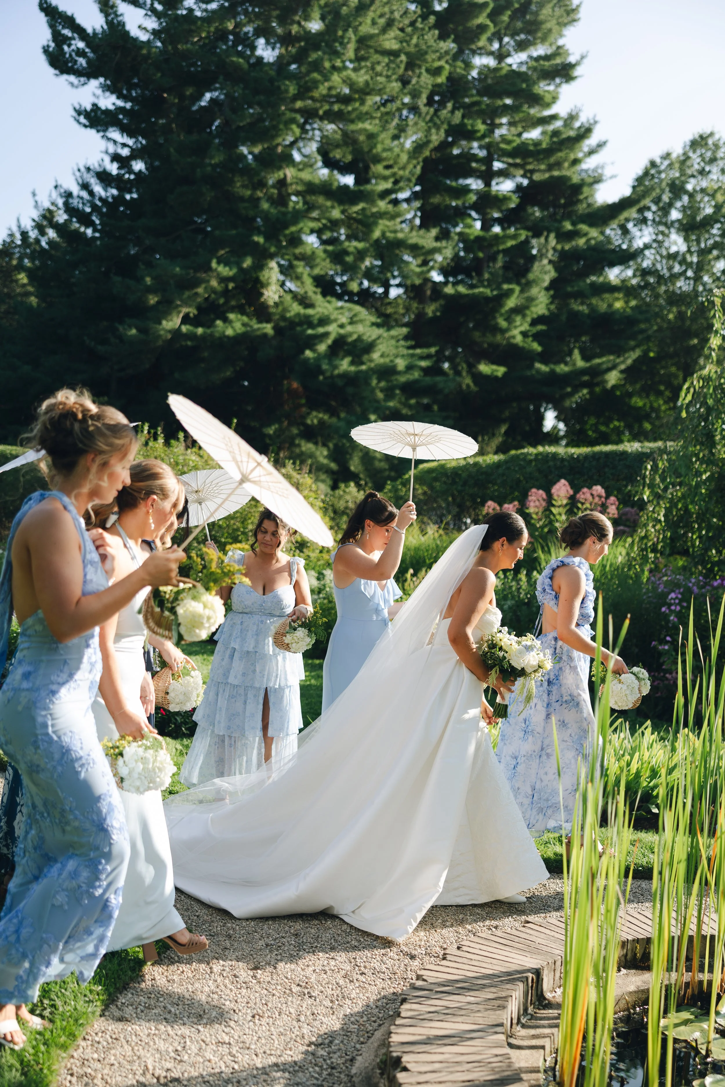 A wedding ceremony taking place outdoors in a garden, with bridesmaids holding white umbrellas and wearing light blue and white dresses, and the bride in a white gown with a long train, surrounded by greenery and tall trees.
