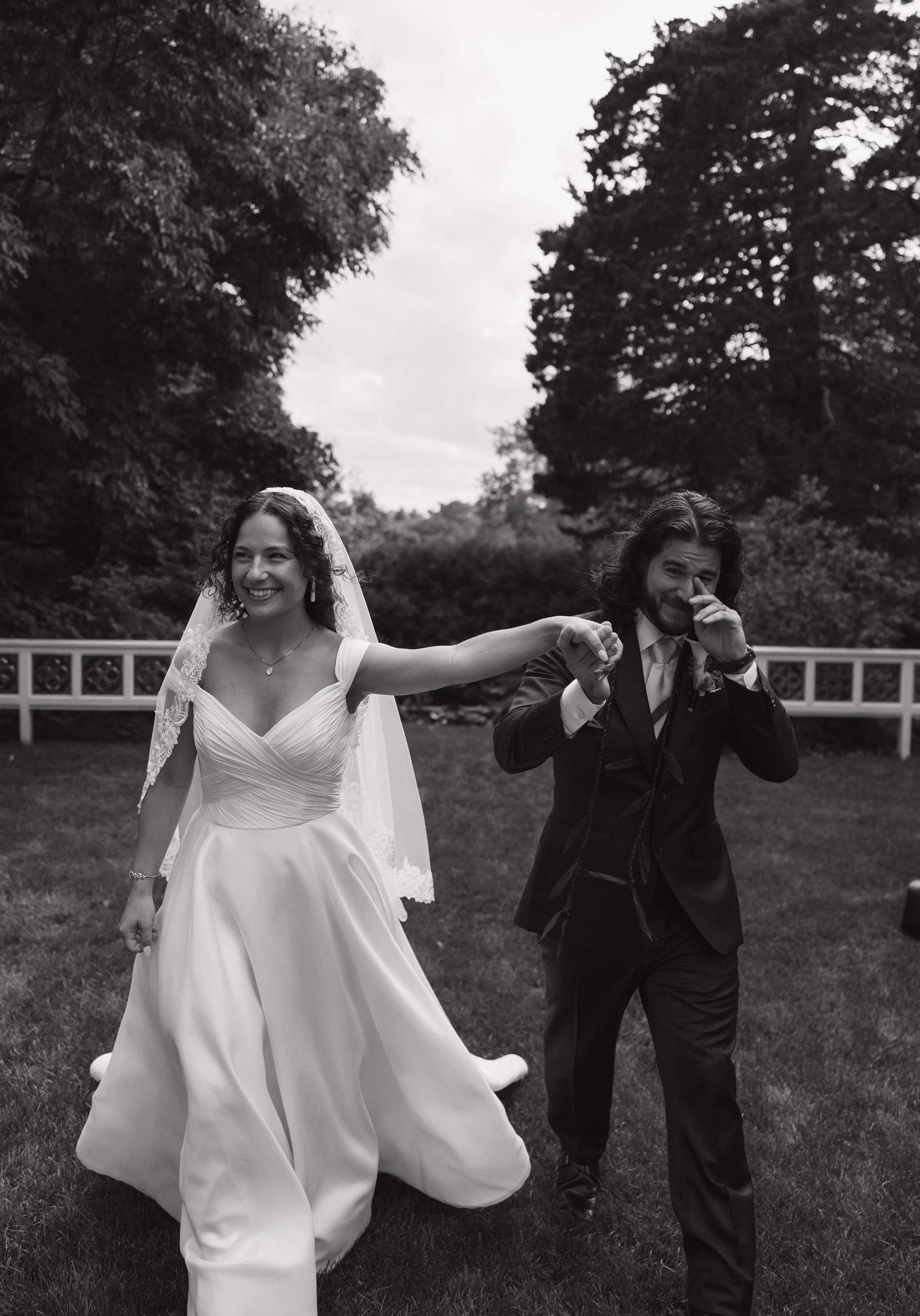 A bride and groom are dancing outdoors on a grassy area, holding hands and smiling, with tall trees and a white fence in the background.
