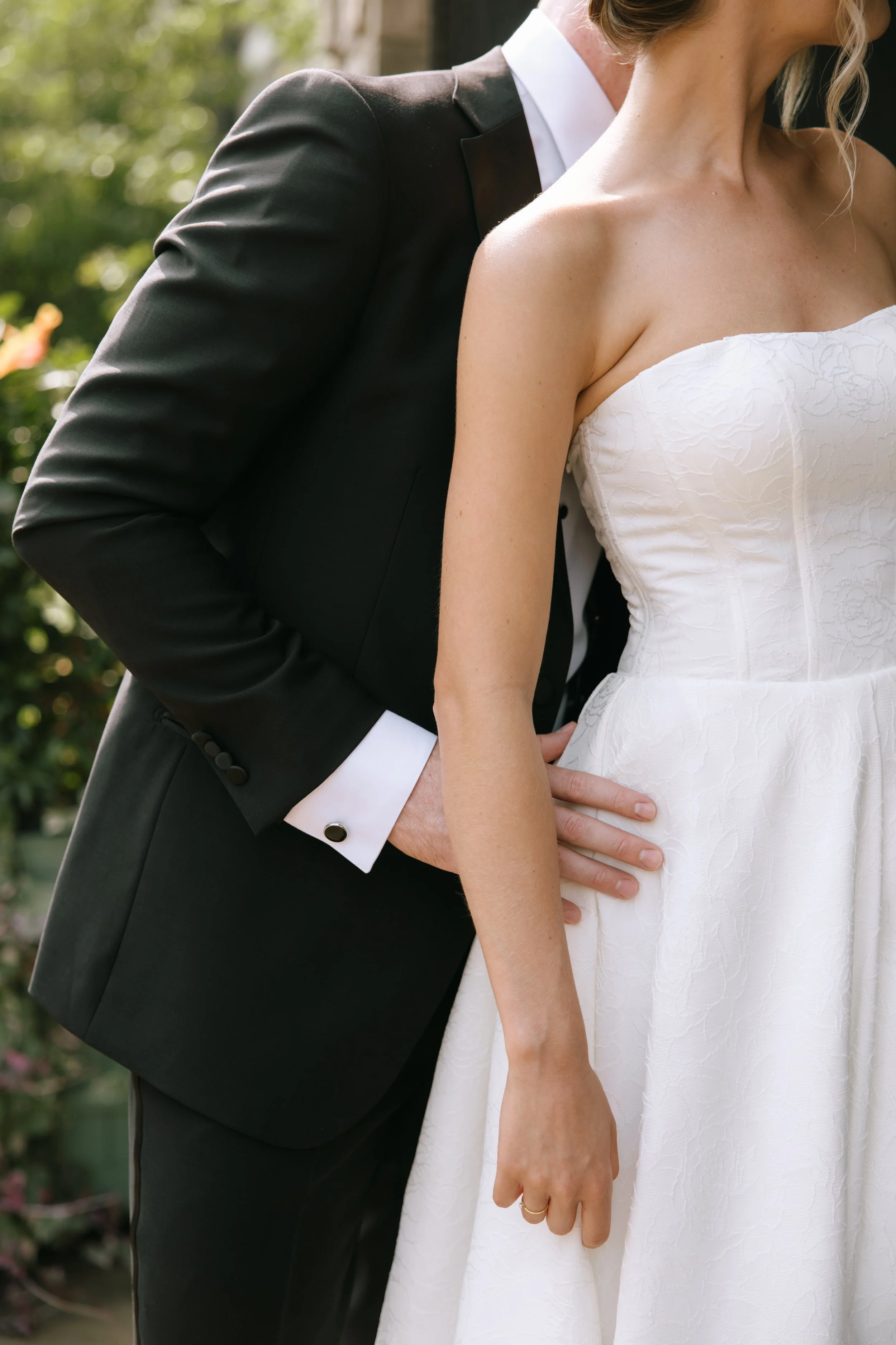 Close-up of a couple dressed in wedding attire sharing an intimate moment outdoors, with the focus on their torsos and the woman's arm gently resting on her waist.