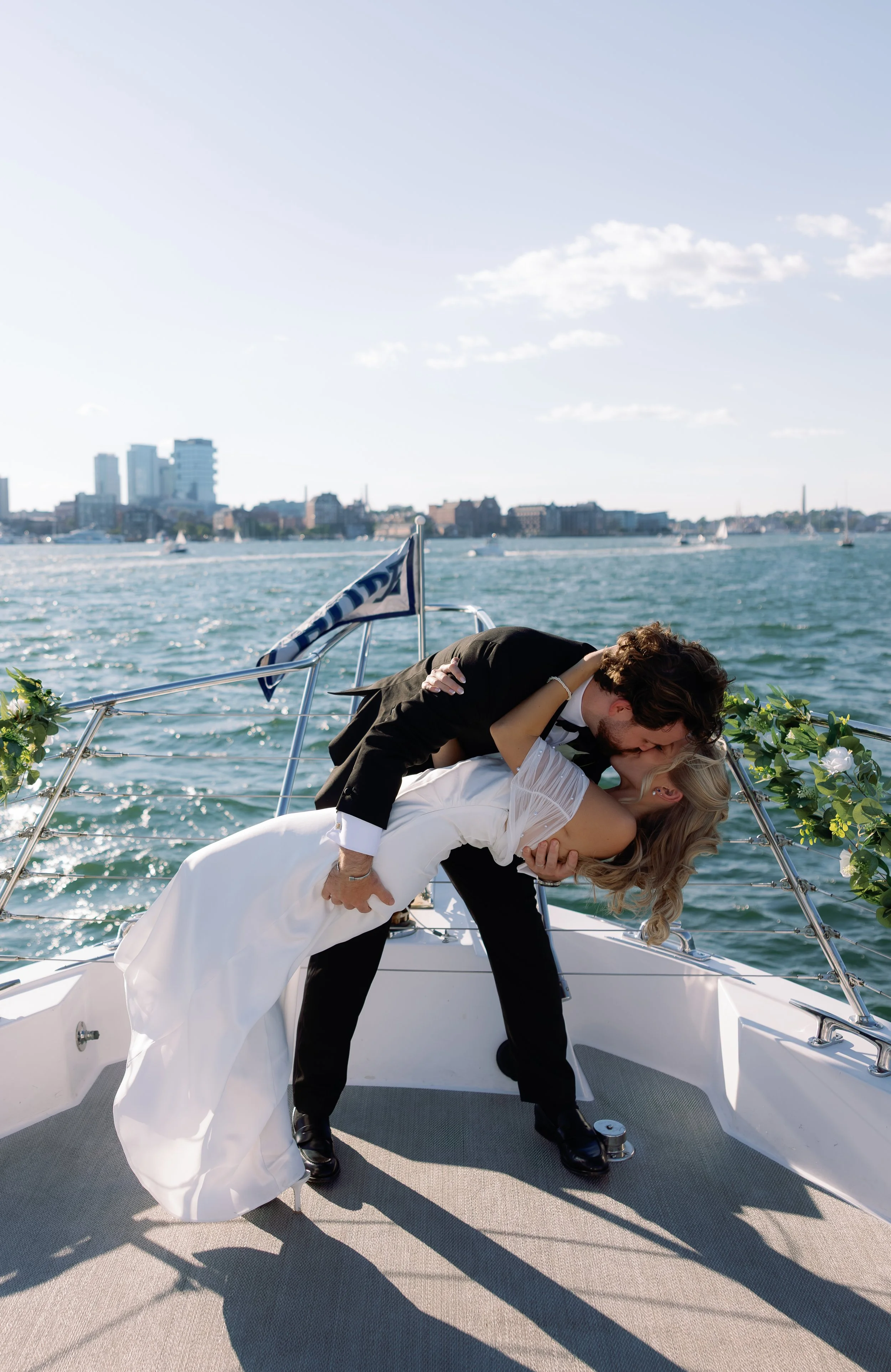 A couple in wedding attire sharing a kiss on a boat with a city skyline and water with sailboats in the background.