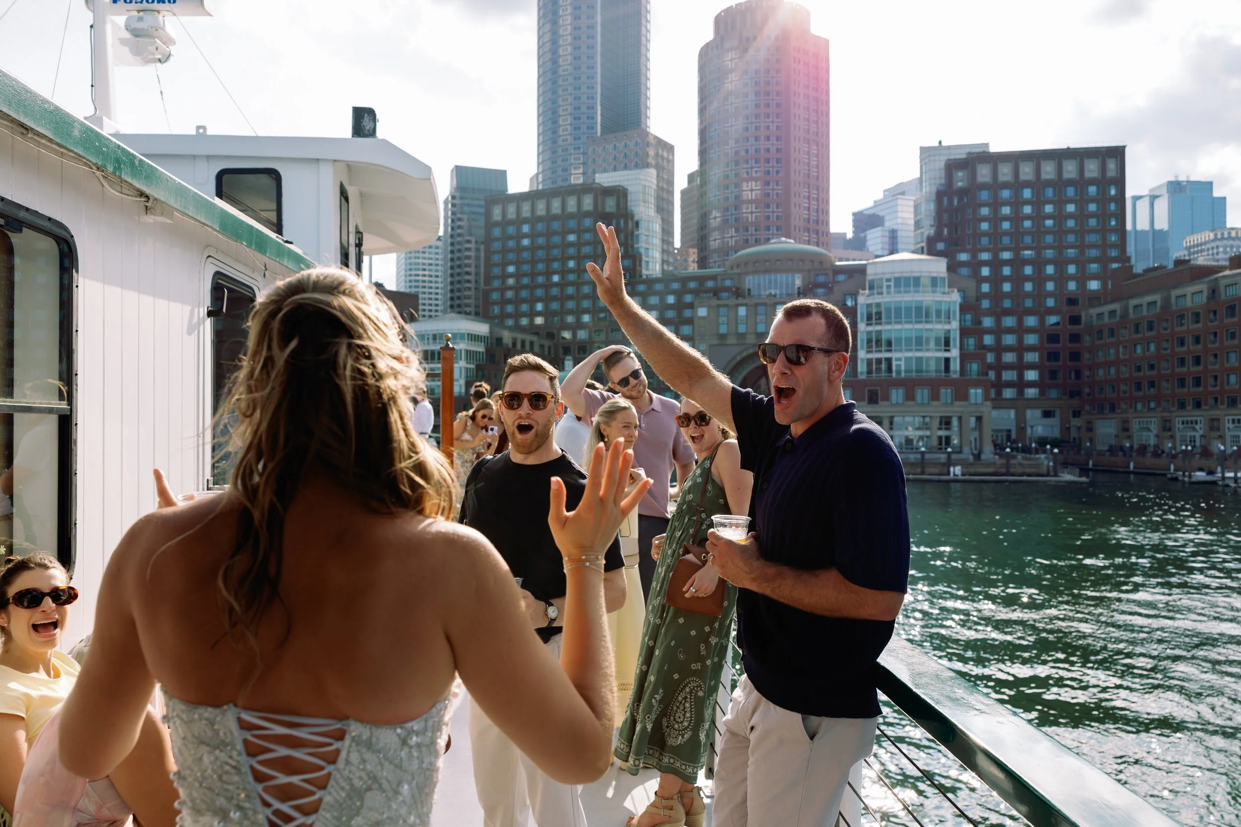 People socializing and enjoying drinks on a boat against a city skyline with tall buildings and a river.