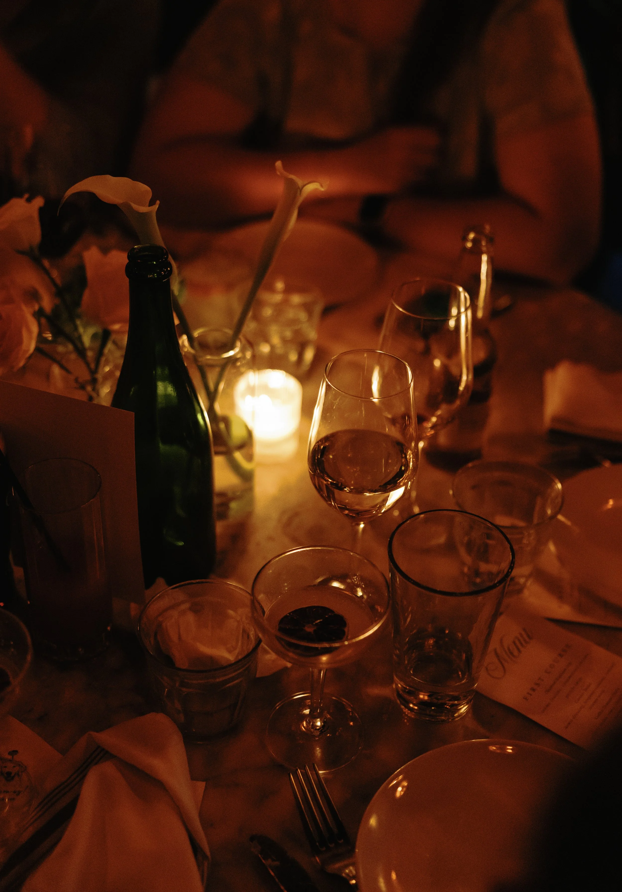A dimly lit table set for a dinner, with various glasses, a wine or champagne bottle, a candle, a vase with flowers, and some menus or papers.