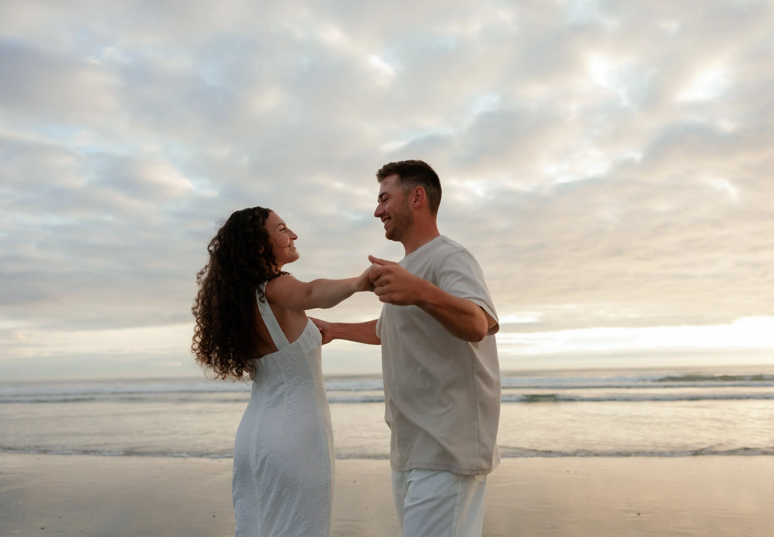 A couple dancing on the beach during sunset, holding hands and looking at each other, with clouds in the sky and ocean waves in the background.