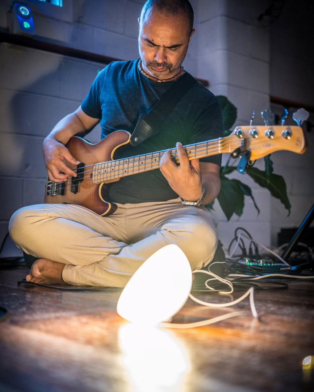 A man sitting cross-legged on the floor playing an electric bass guitar, with a small white lamp on the floor nearby.