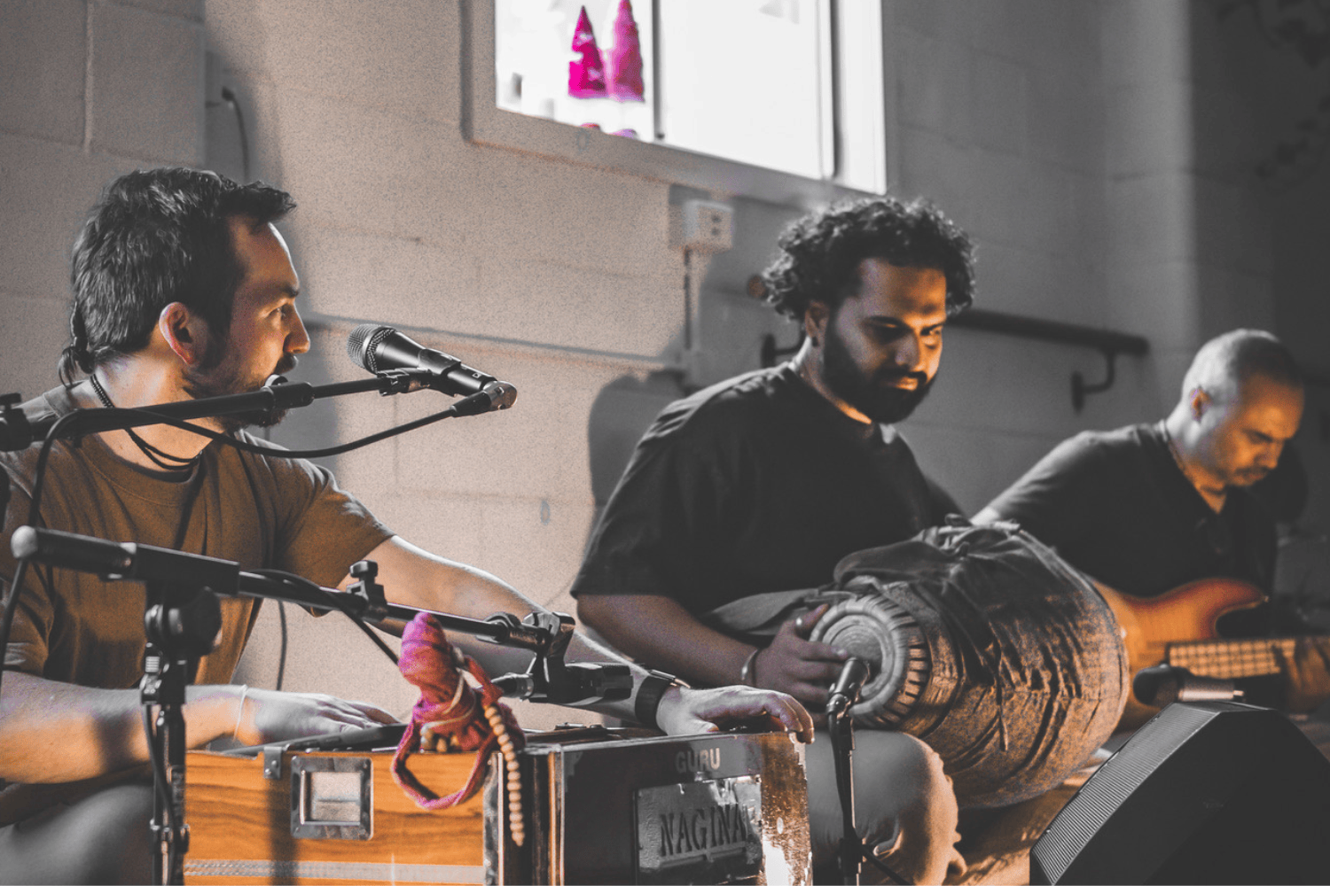 Three musicians sitting on the floor, playing instruments, one with a keyboard and microphone, another with a percussion instrument, and the third with a guitar, in a room with minimal decor and a window with pink and purple art.