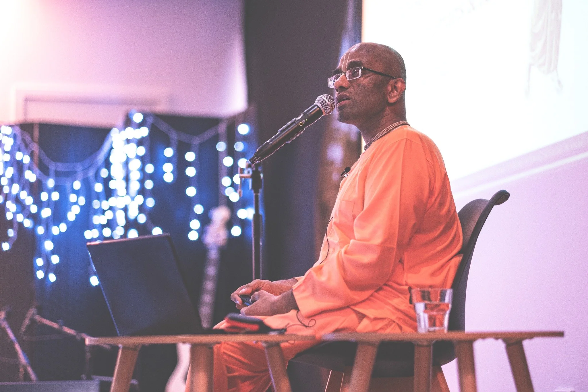 Three musicians sitting on the floor, playing instruments, one with a keyboard and microphone, another with a percussion instrument, and the third with a guitar, in a room with minimal decor and a window with pink and purple art.