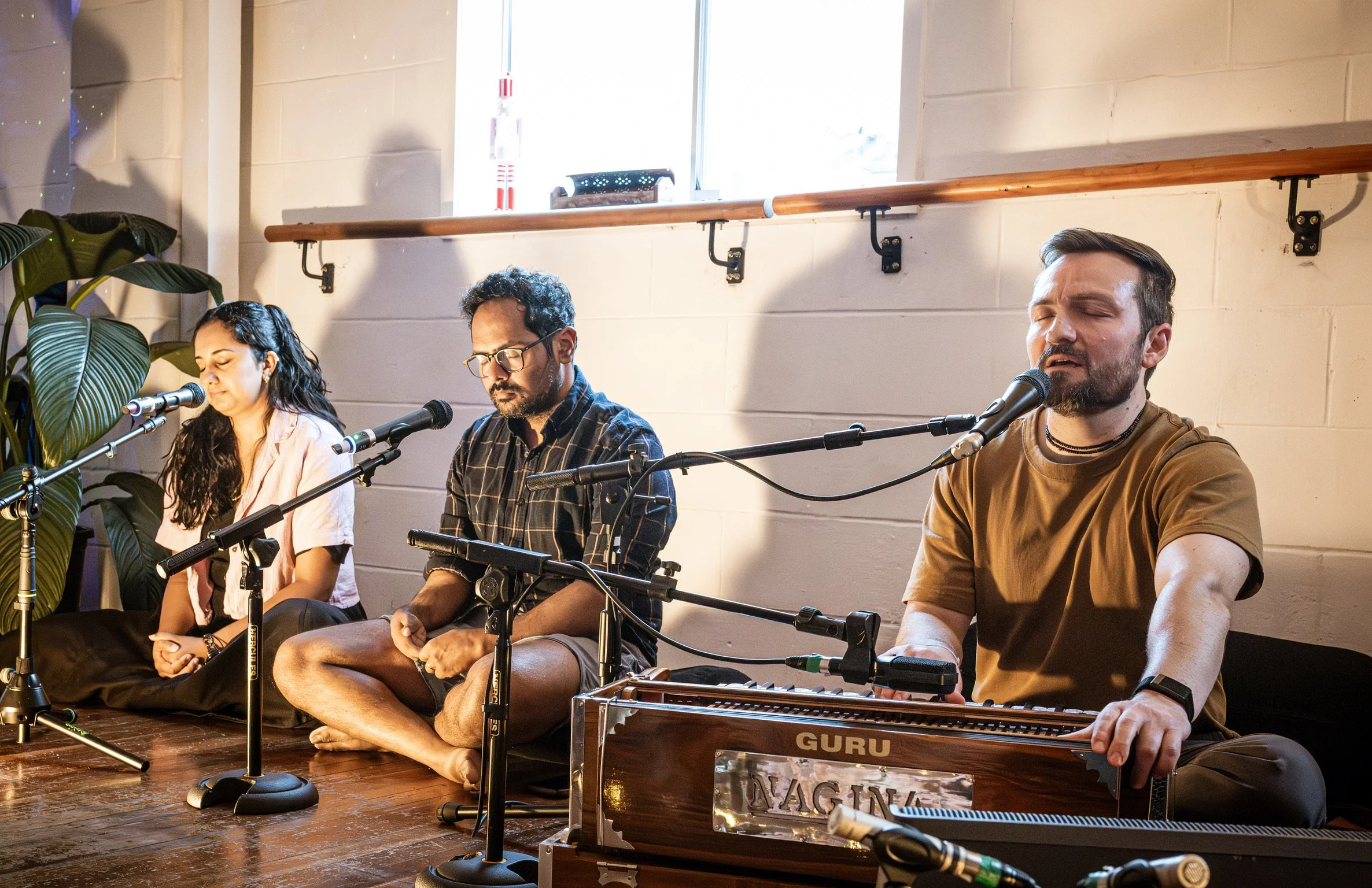 Three musicians sitting on the floor, singing and playing instruments. The woman on the left has long dark hair, the man in the middle has glasses and curly hair, and the man on the right has a beard and is playing a keyboard labeled 'GURU.'