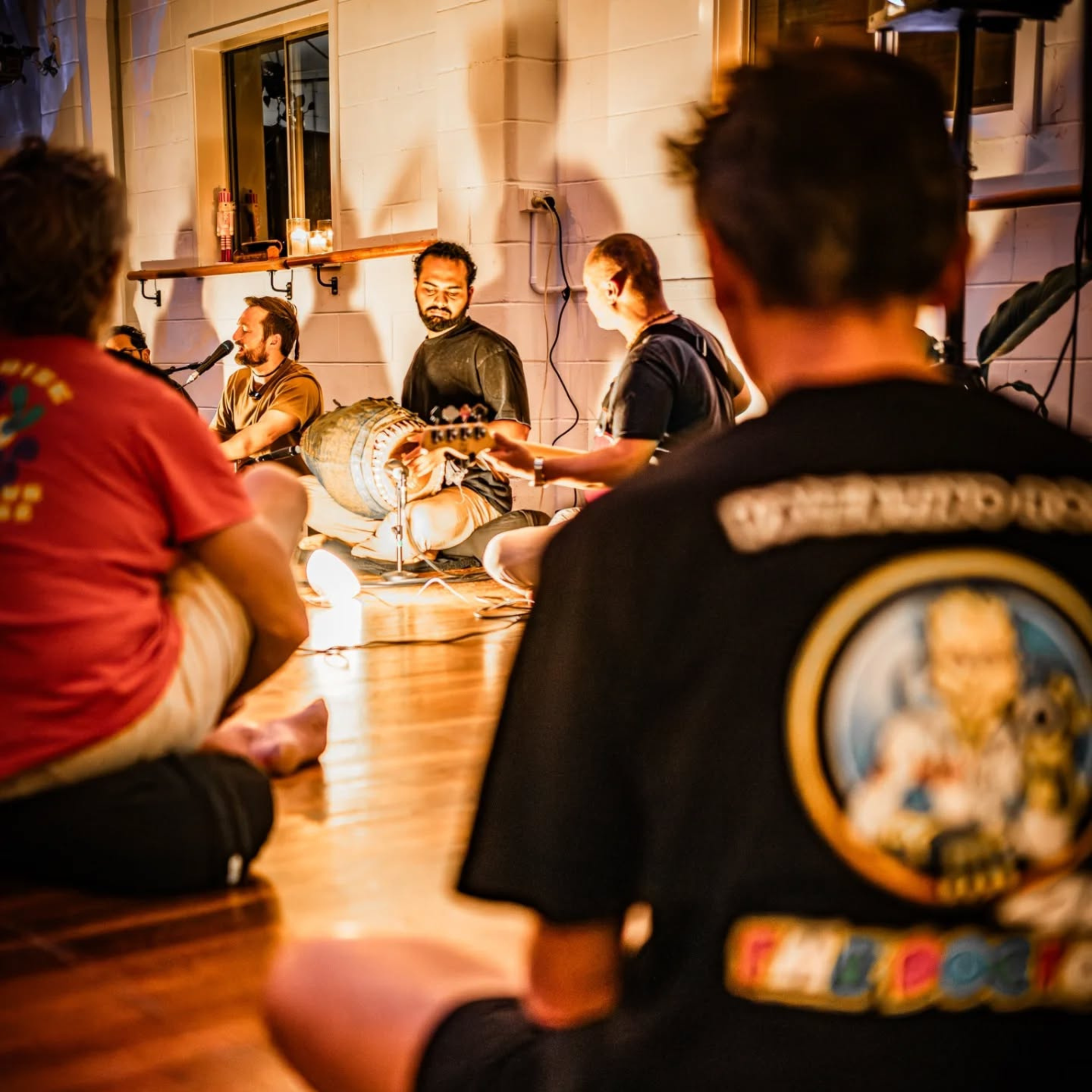 Group of people sitting on the floor in a cozy indoor setting, engaging in a musical performance with instruments and microphones, warm lighting, casual atmosphere.