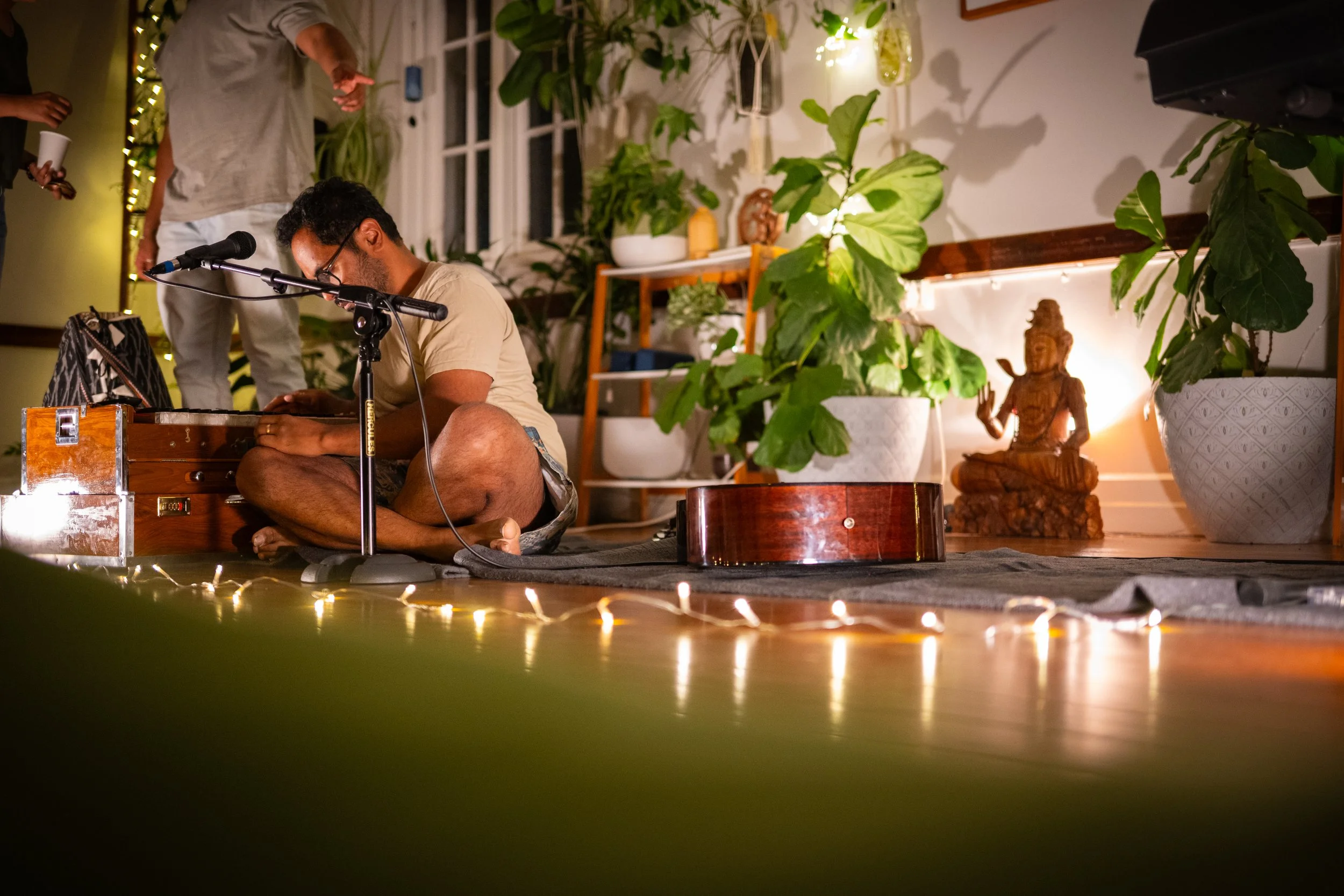 A man sitting cross-legged on the floor near a microphone, playing a harmonium with a guitar nearby, in a cozy room decorated with plants, a Buddha statue, and string lights.