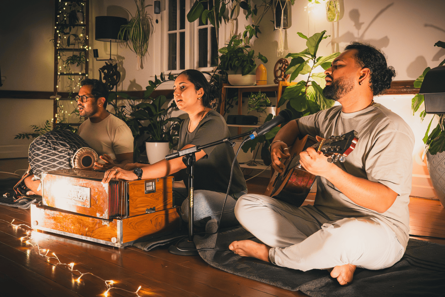Three people are sitting on the floor in a cozy room, engaging in a musical performance. They are surrounded by plants and warm lighting, with one person playing a guitar, another playing a hand drum, and the third singing into a microphone.
