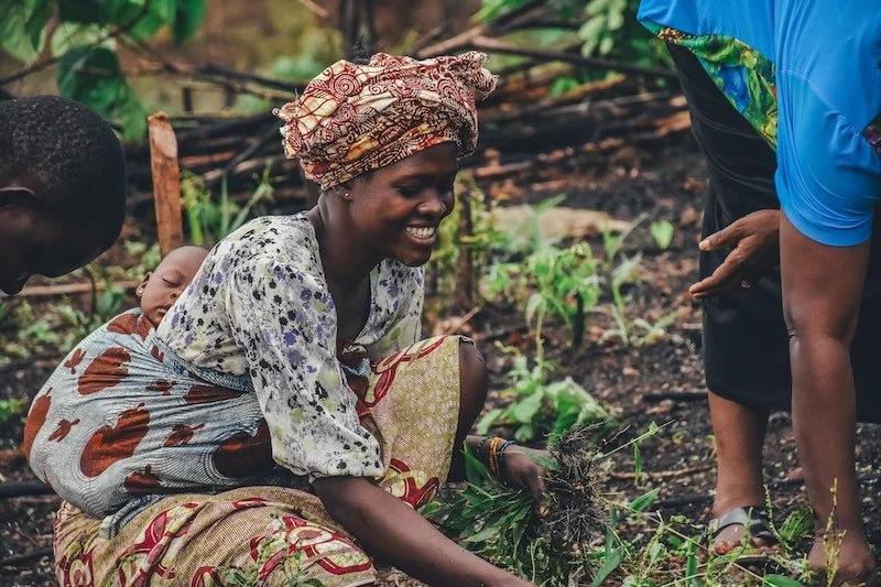 A woman kneeling in a garden with young plants, smiling and holding a plant with roots, with a child on her back. Another person is standing nearby. The garden is lush and green.