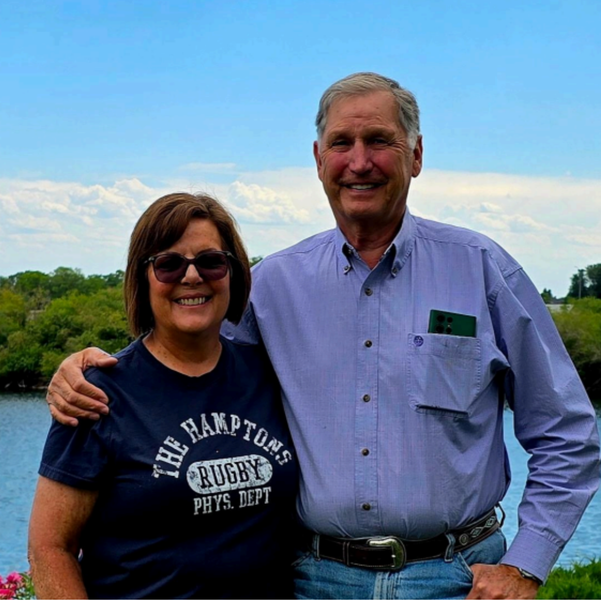 A smiling woman wearing sunglasses and a navy t-shirt with white text, standing outdoors with a smiling man in a light blue button-up shirt, near a body of water and green trees under a partly cloudy sky.