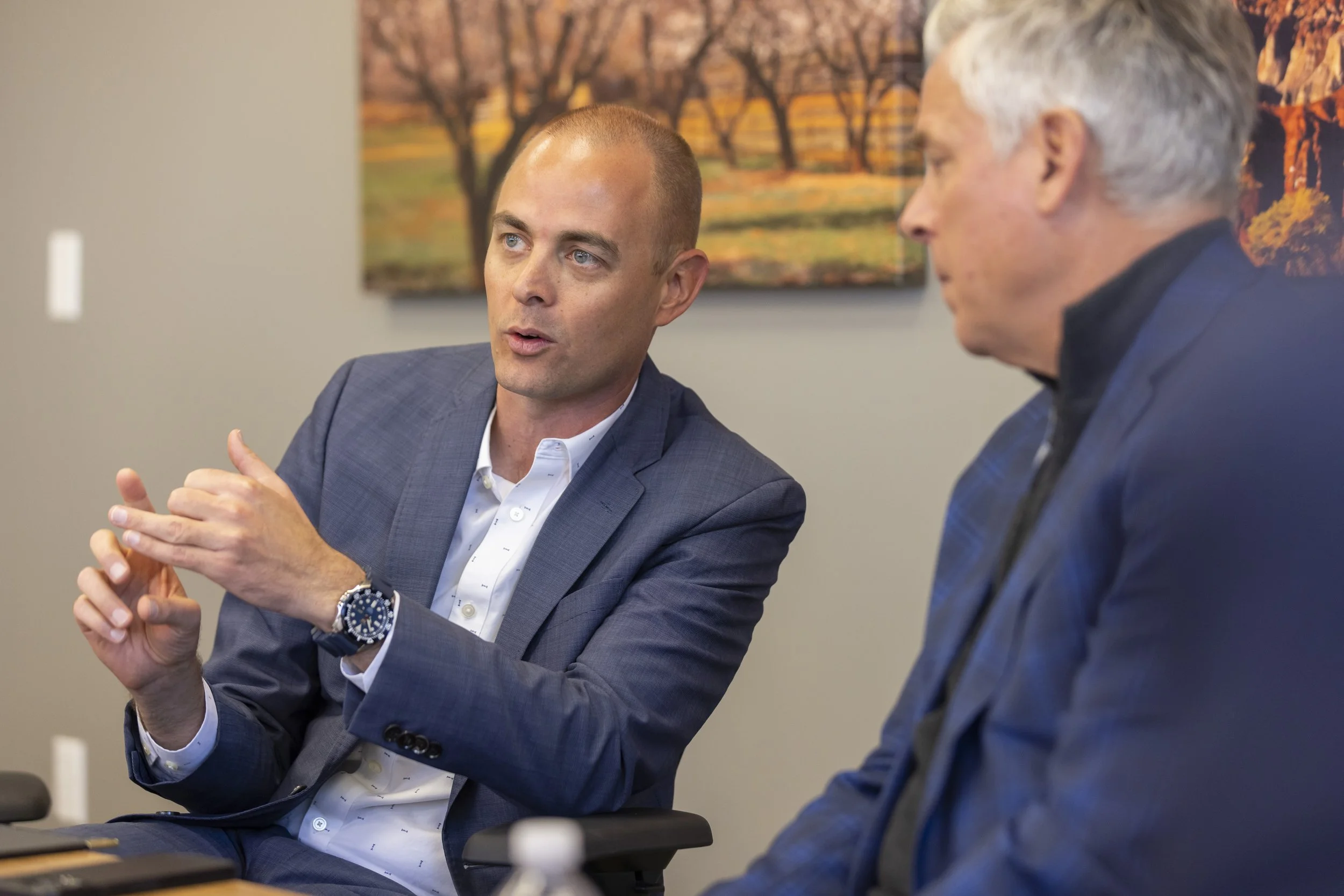 Two men in business suits engaged in a discussion, sitting at a table in an office with a painting of autumn trees in the background.