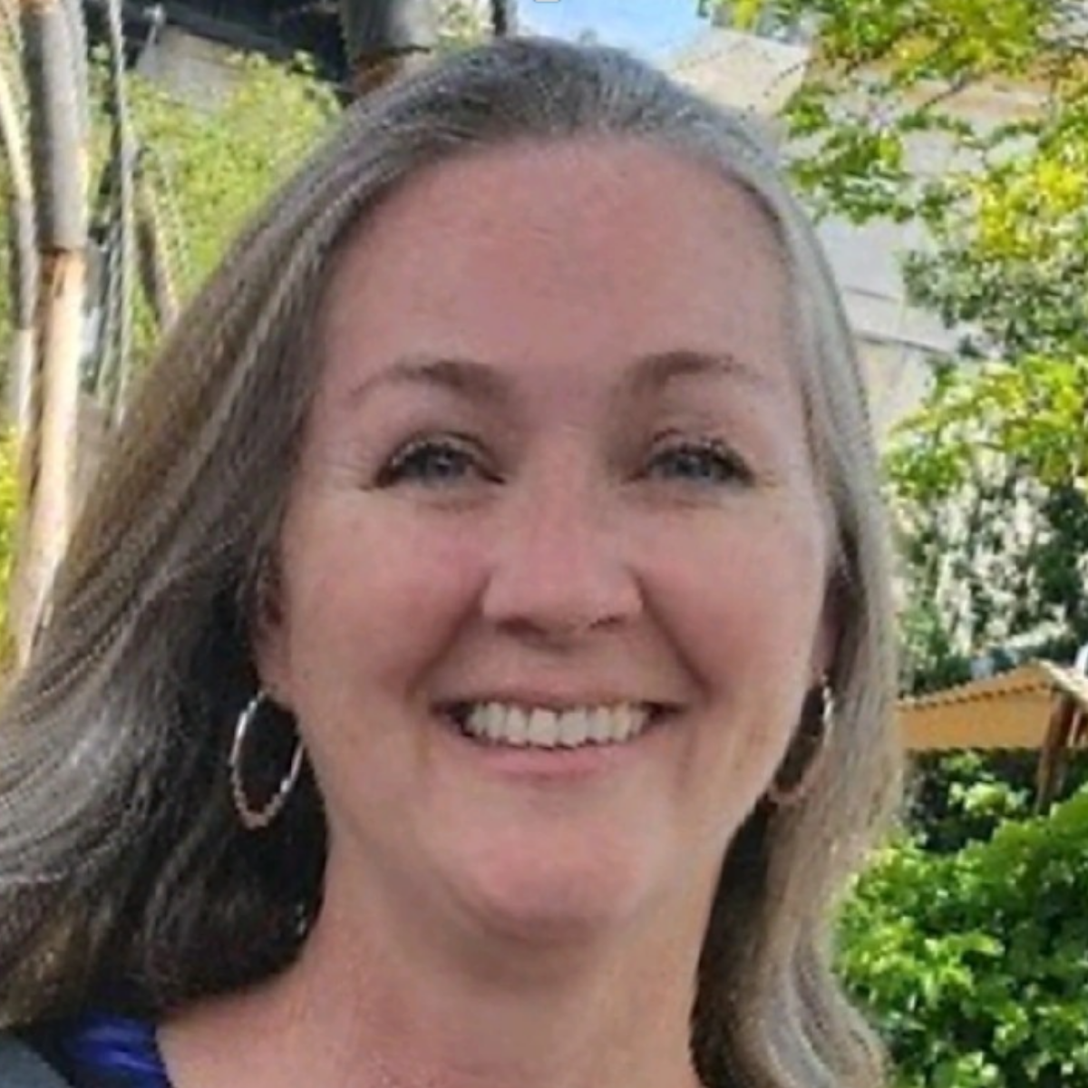 A smiling woman with long blonde hair and hoop earrings, outdoors with trees and a building in the background.