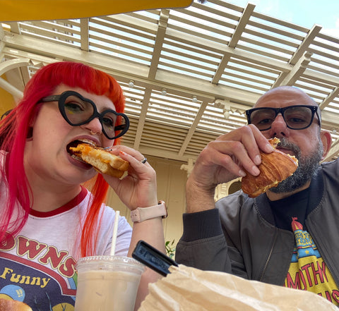 A woman with bright red hair and a man with a beard and glasses eating bread or toast at a table outdoors.