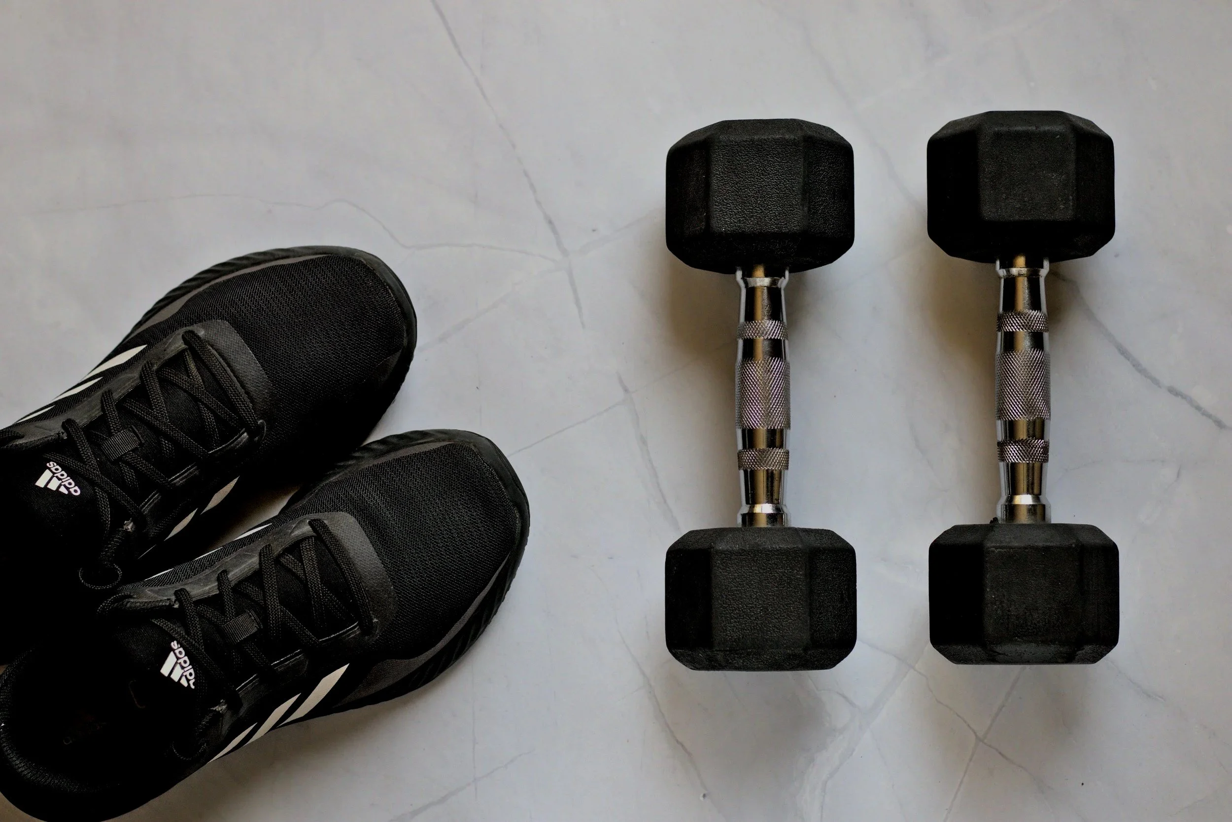 Une paire de chaussures de sport noires, et deux haltères avec des poids en forme de cube sur une surface en marbre