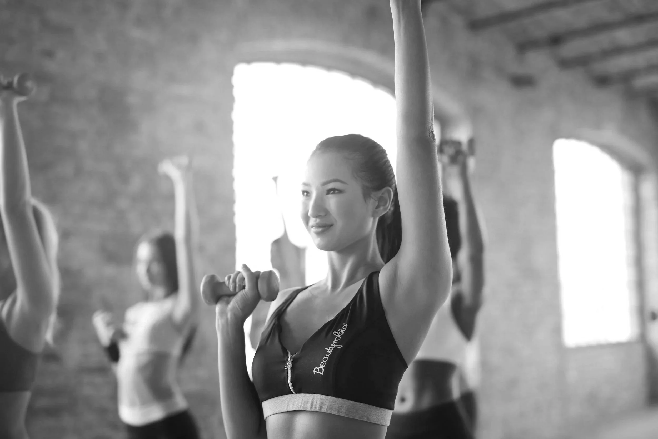 Une femme souriante dans une salle de sport, faisant de l'exercice avec un haltère, entourée d'autres personnes également en train de s'entraîner.