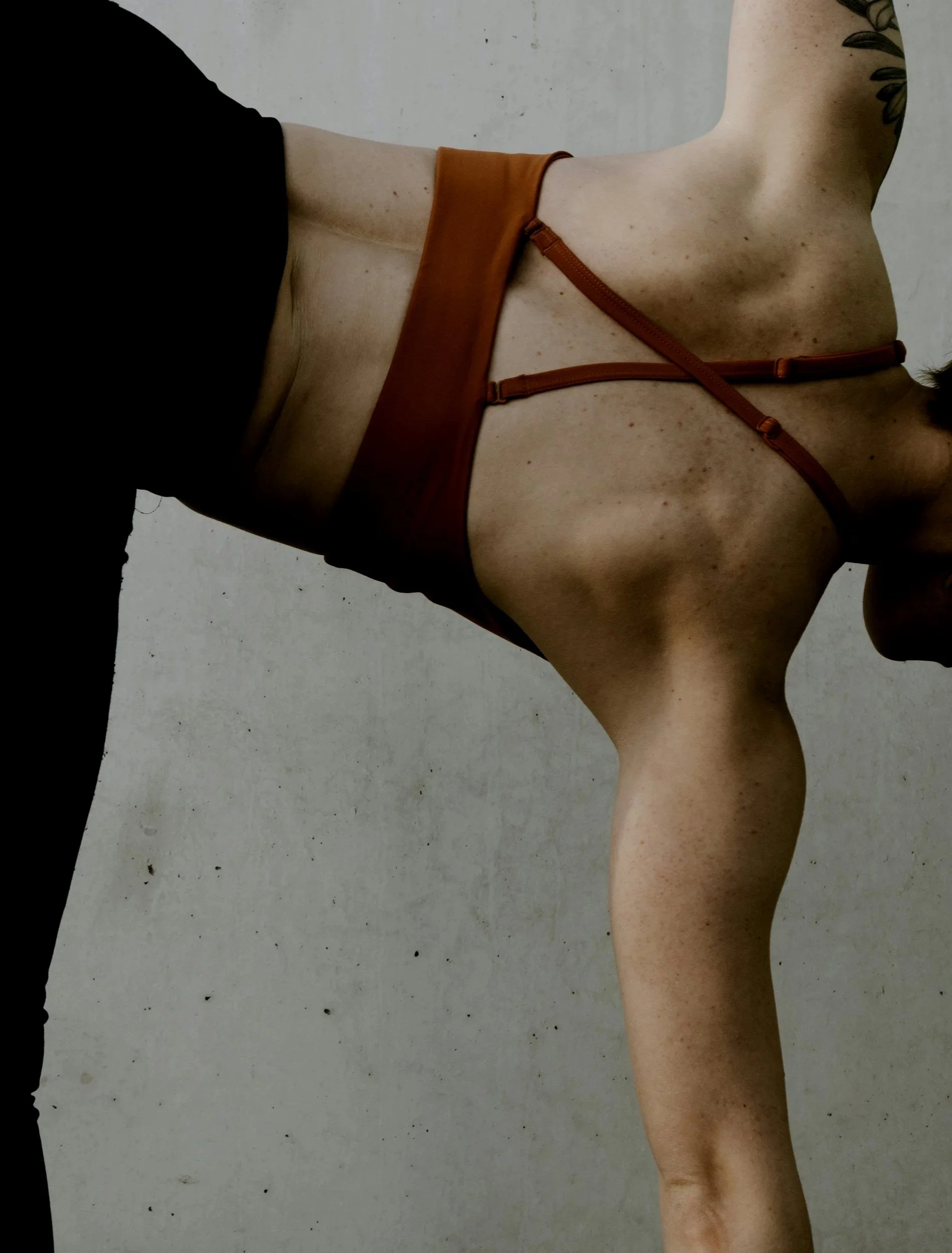 Une femme portant un chemisier en cuir marron, avec des épaules et une poitrine visibles, debout contre un mur gris.