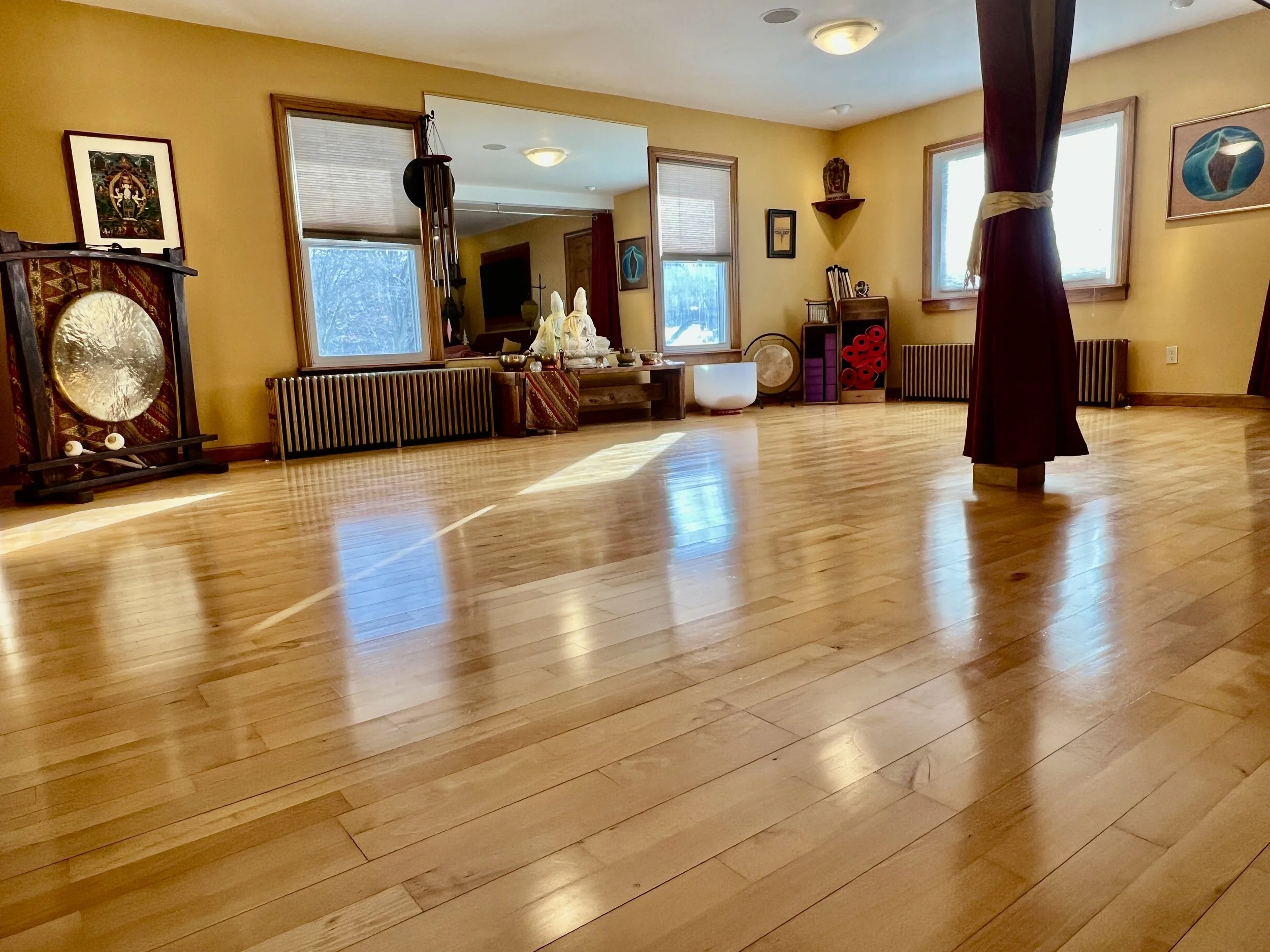A spacious room with polished wooden floors, golden colored walls, large windows, and various decorative items, including a gong, art pieces, and a curtain tied to a pillar.