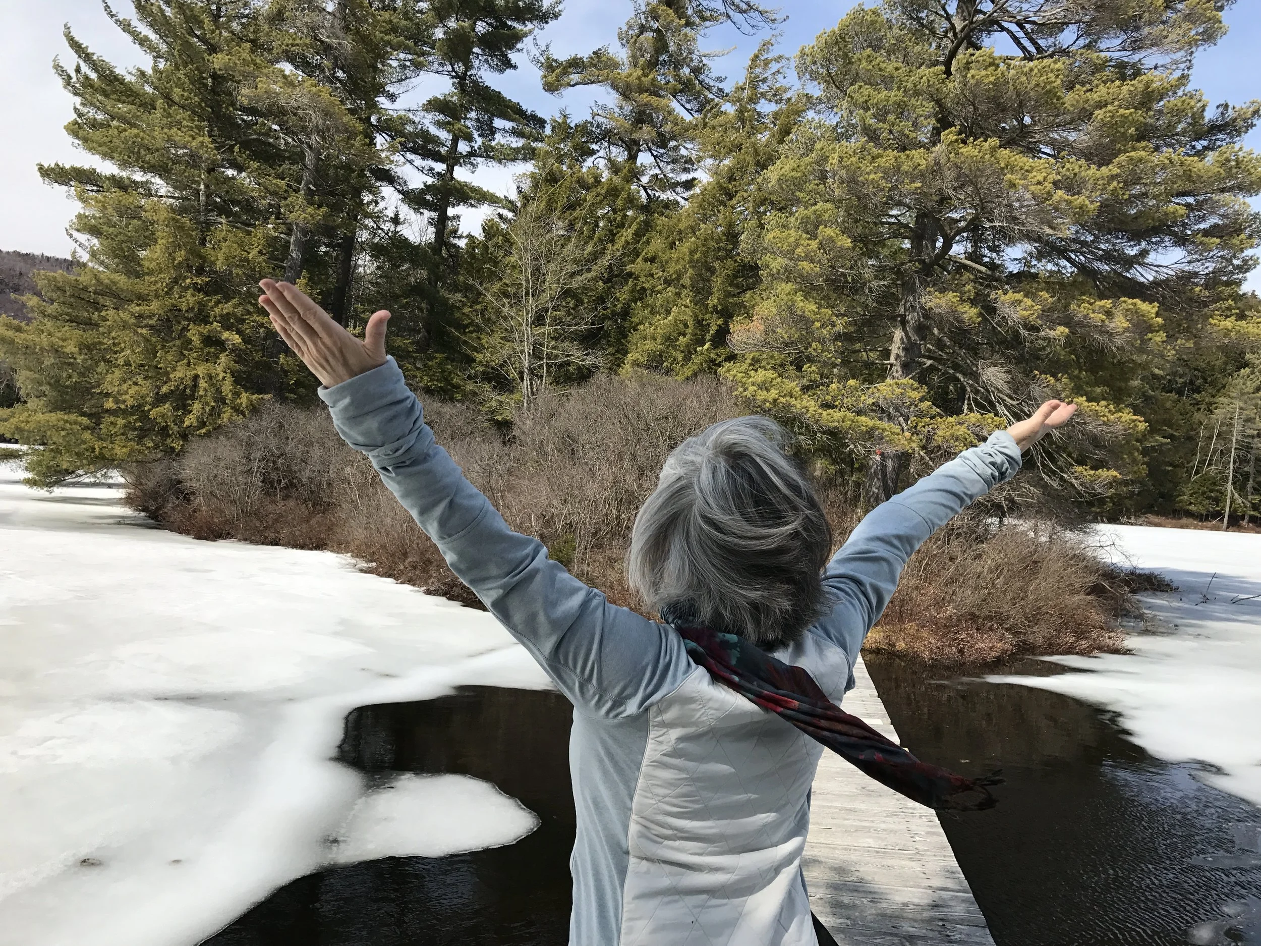 Older woman with gray hair, feeling the freedom, standing outdoors with arms outstretched in front of a partially frozen river and tall evergreen trees in the Adirondacks.