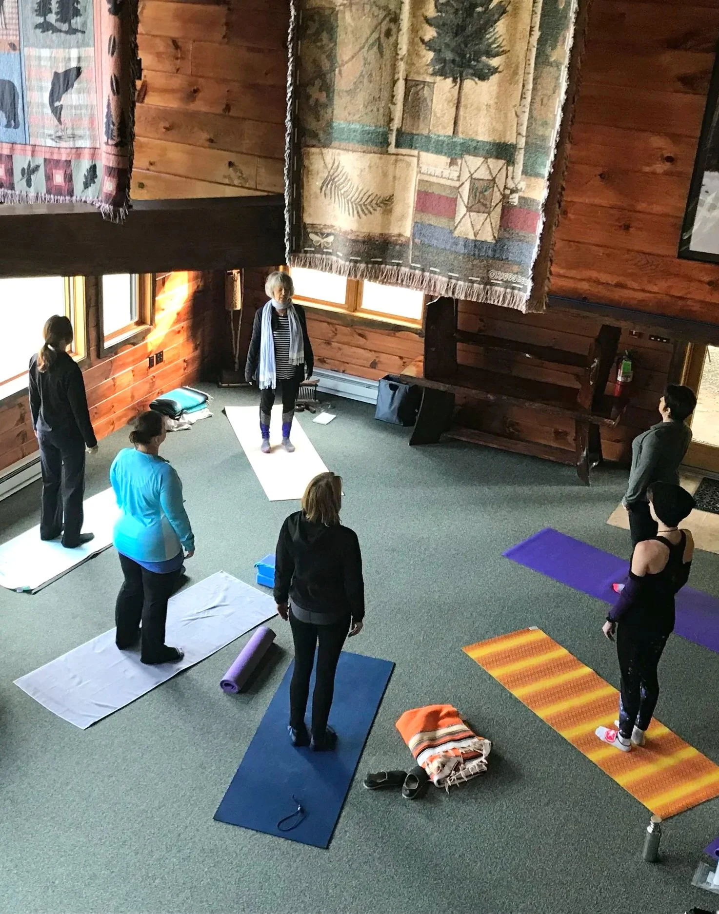 A group of people in a yoga class in a rustic wooden room, standing on yoga mats, with Mary Bartel leading the session.
